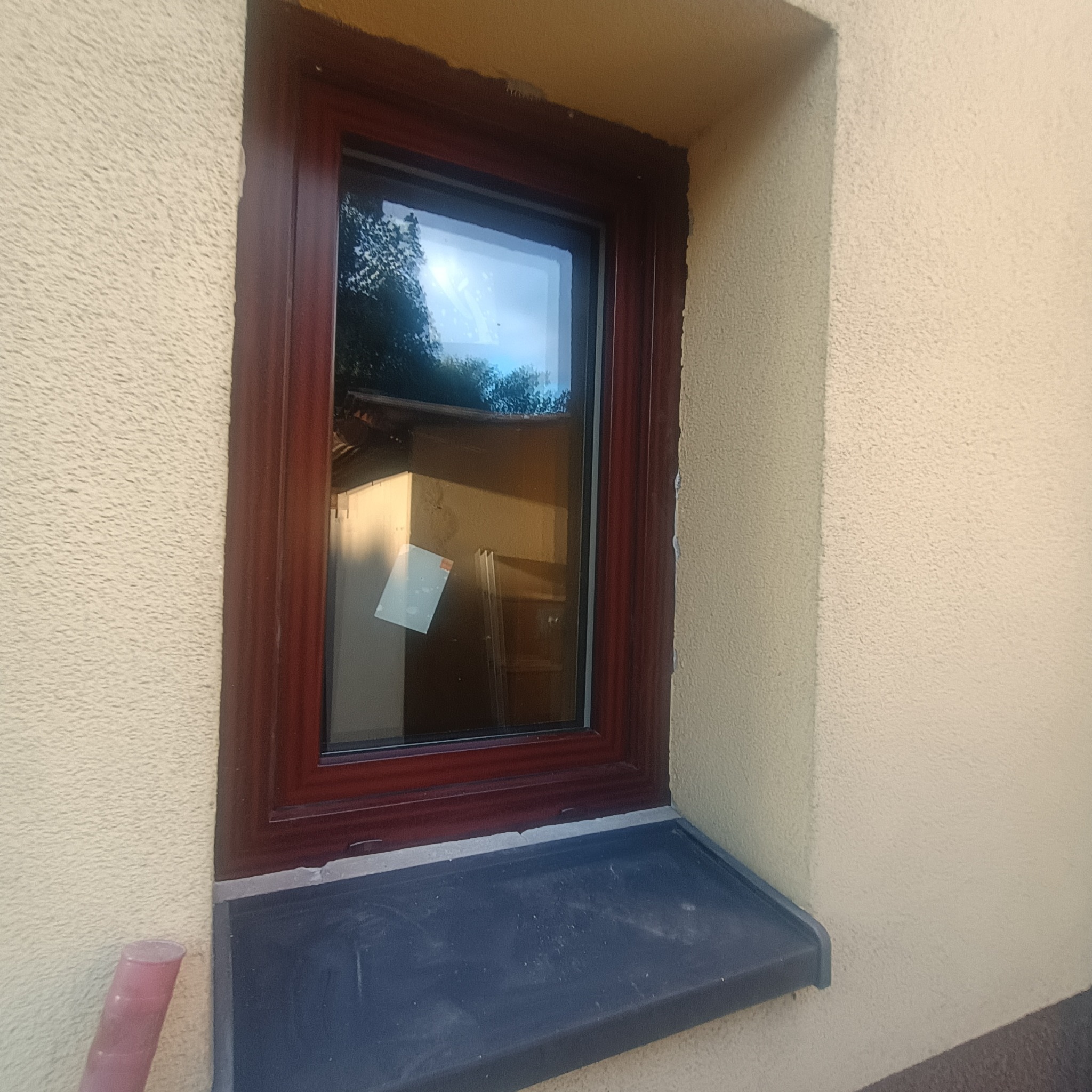 Zamontowane okno z brązową ramą w elewacji budynku o jasnej, ziarnistej strukturze. Widoczny parapet i odblask otoczenia w szybie.