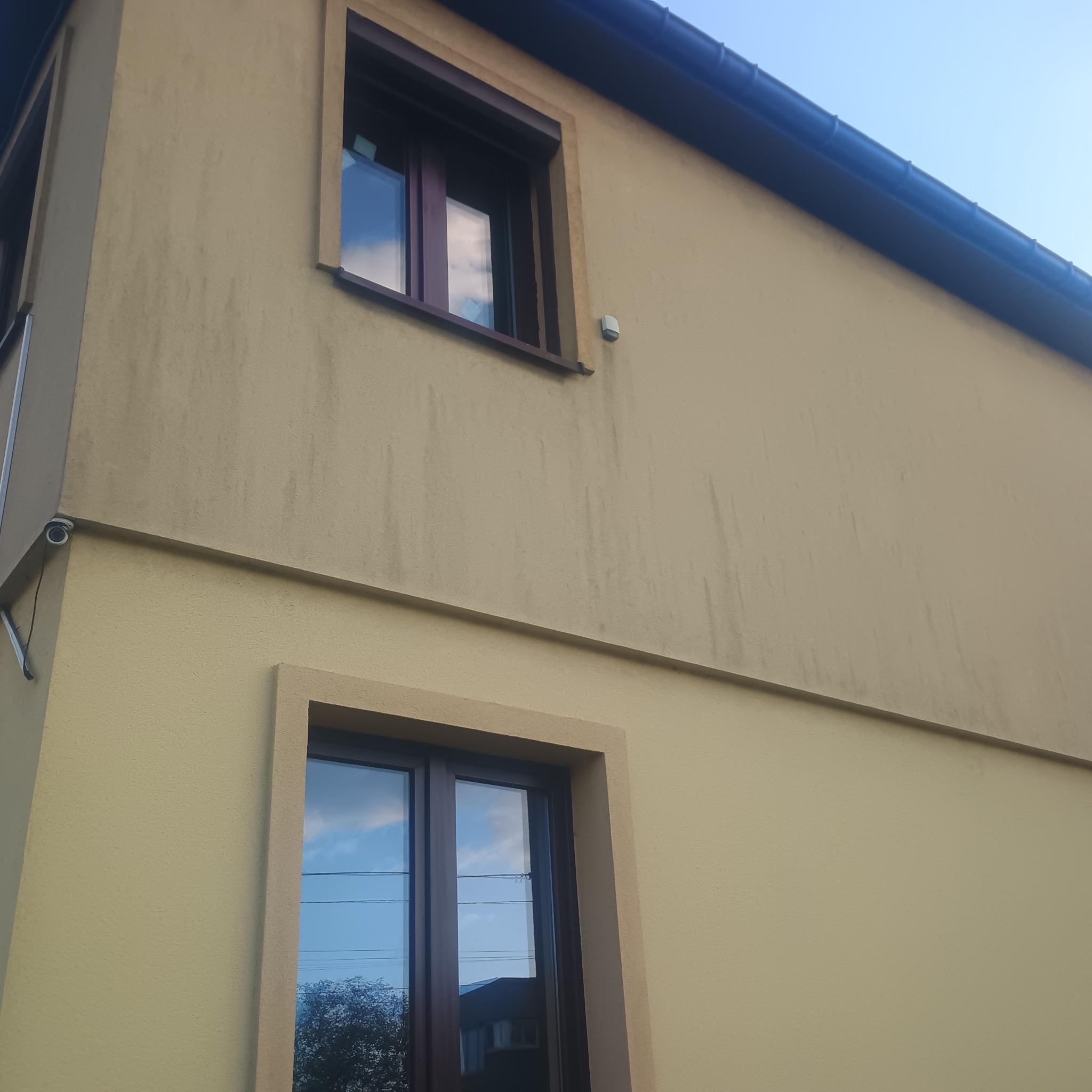 Fragment fasady budynku z dwoma oknami z brązowymi ramami. W szybach odbija się niebo. Widoczny fragment dachu z rynną i kamera monitoringu.