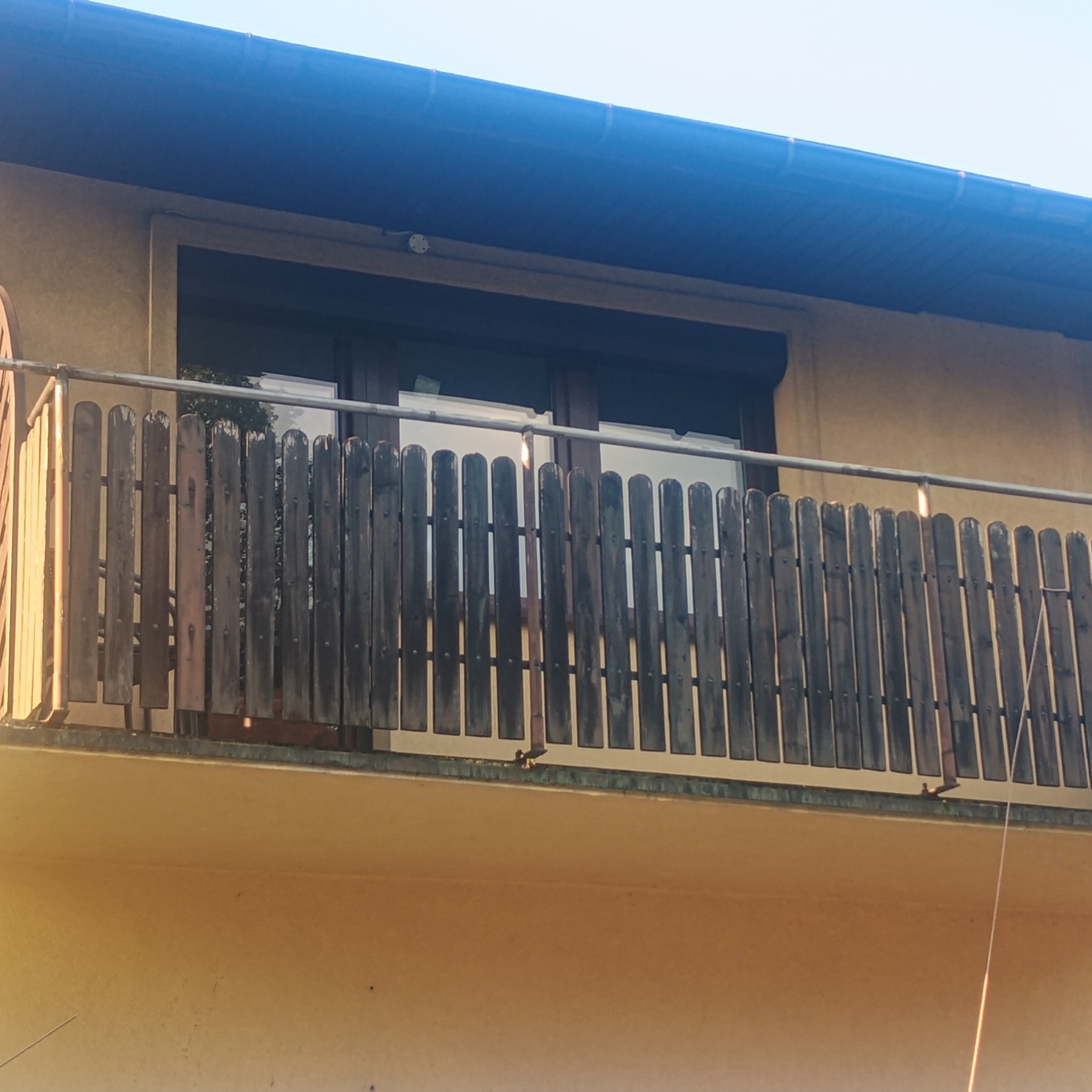 Balkon z drewnianą balustradą, widok z dołu. Na drugim planie okno z roletą. Elementy mocujące balustradę wykonane z metalu. Budynek o żółtej elewacji.