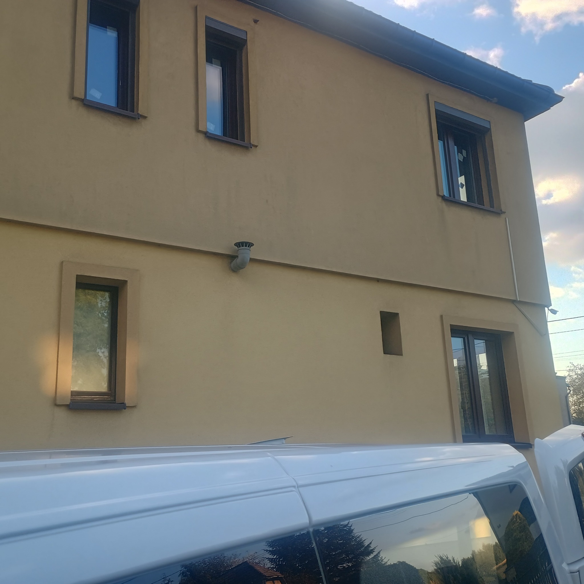 Elewacja budynku z oknami o brązowych ramach, częściowo zasłonięta przez biały samochód dostawczy. Widoczne okna na różnych poziomach budynku.