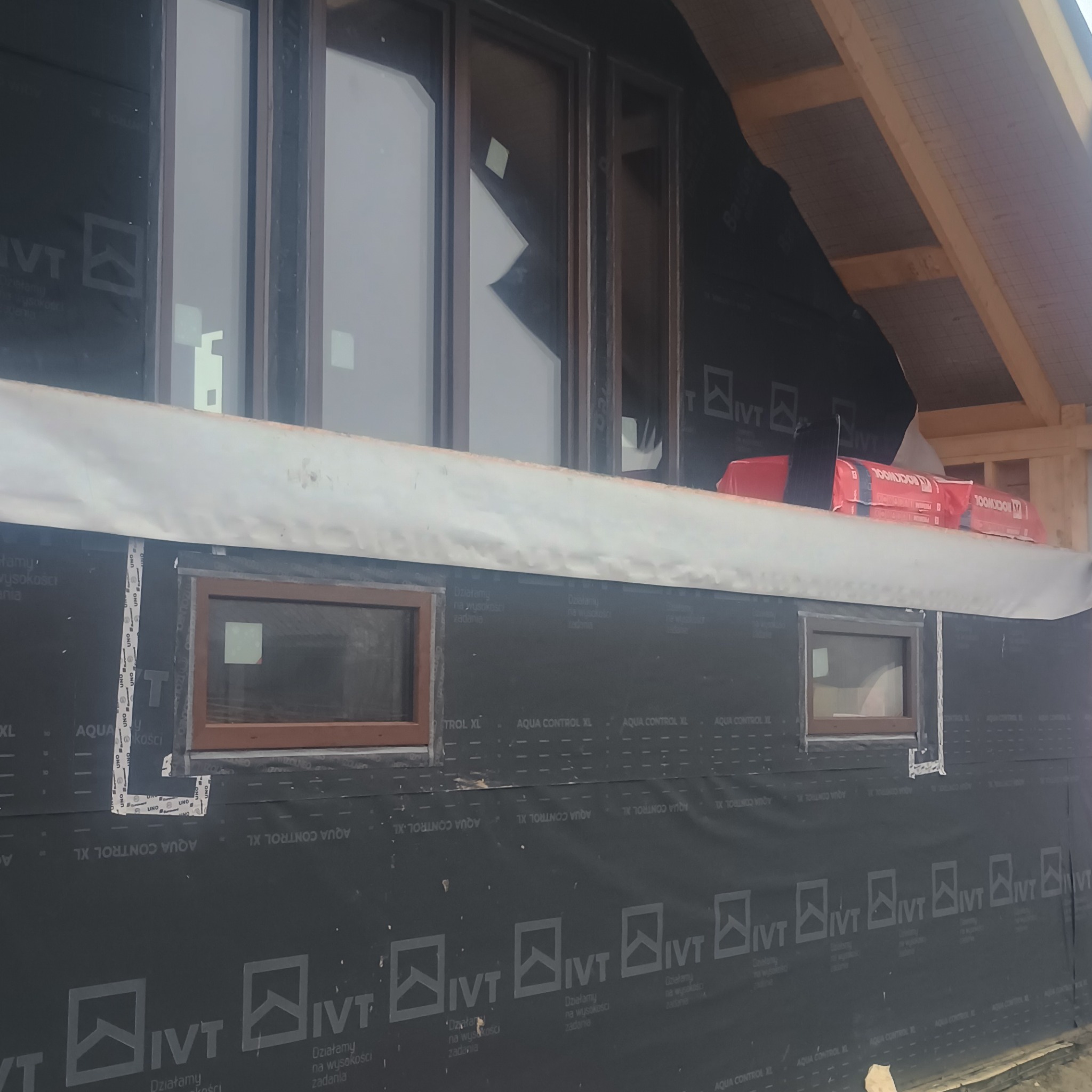 Montaż okien w nowym budynku, widoczne okna z brązowymi ramami i czarna membrana na ścianie. Częściowo widoczna konstrukcja dachu.