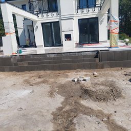 Marek Wieczorkiewicz - Budowa tarasu z kostki brukowej przy nowoczesnym domu. Widoczne ułożone stopnie z kostki, przygotowany teren i materiały budowlane. Słupy osłonięte folią.