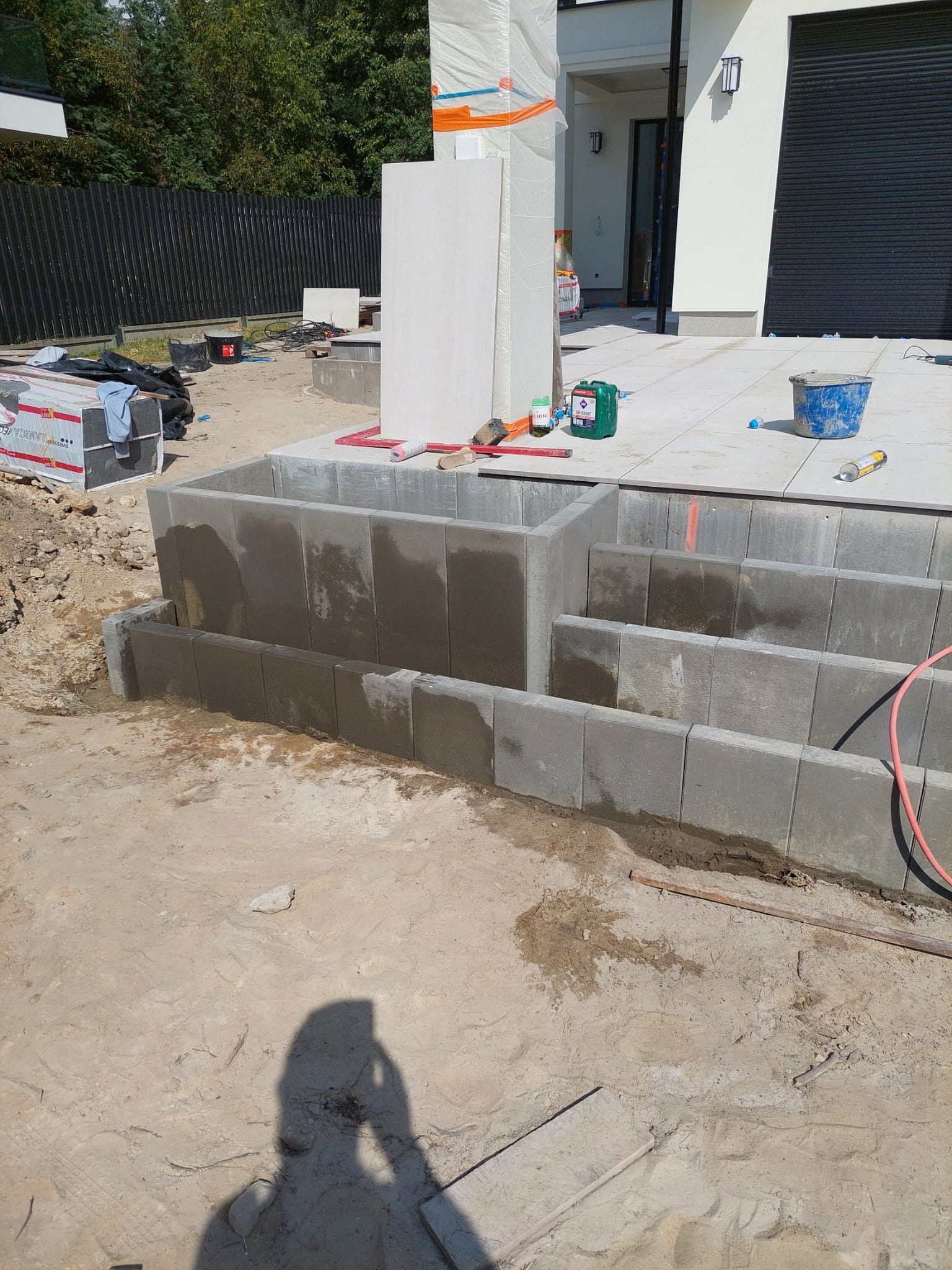 Budowa schodów zewnętrznych z betonowych bloczków, widoczne narzędzia i materiały budowlane, teren przygotowany pod dalsze prace.