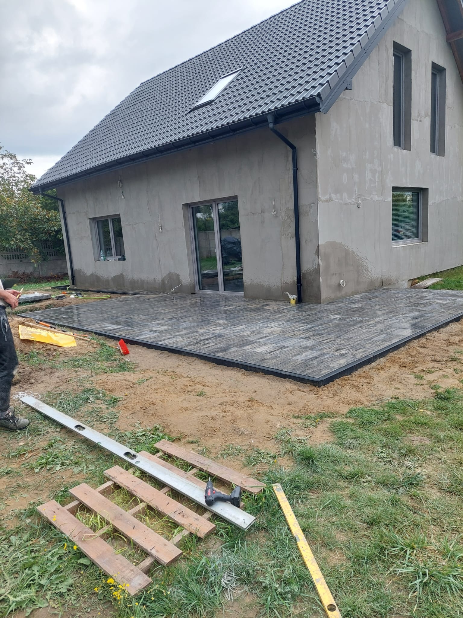 Ułożona kostka brukowa na tarasie przylegającym do surowego domu. Widoczne narzędzia brukarskie i poziomica. Tło stanowi trawa i częściowo wykończona elewacja.