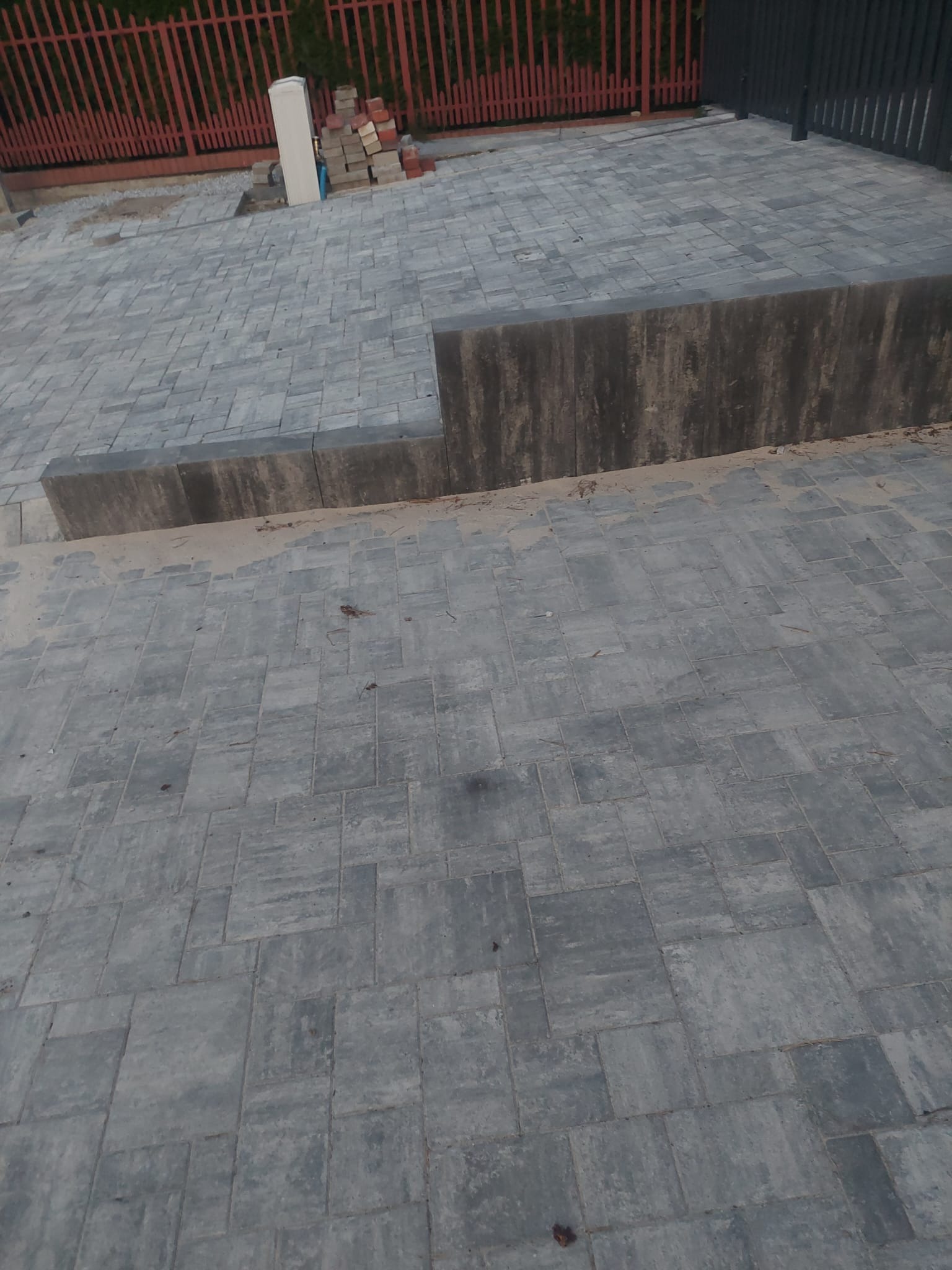 Ułożona szara kostka brukowa tworząca ścieżkę z obrzeżami. Fragment czerwonego ogrodzenia w tle. Widoczne zapasy kostki i materiałów budowlanych.