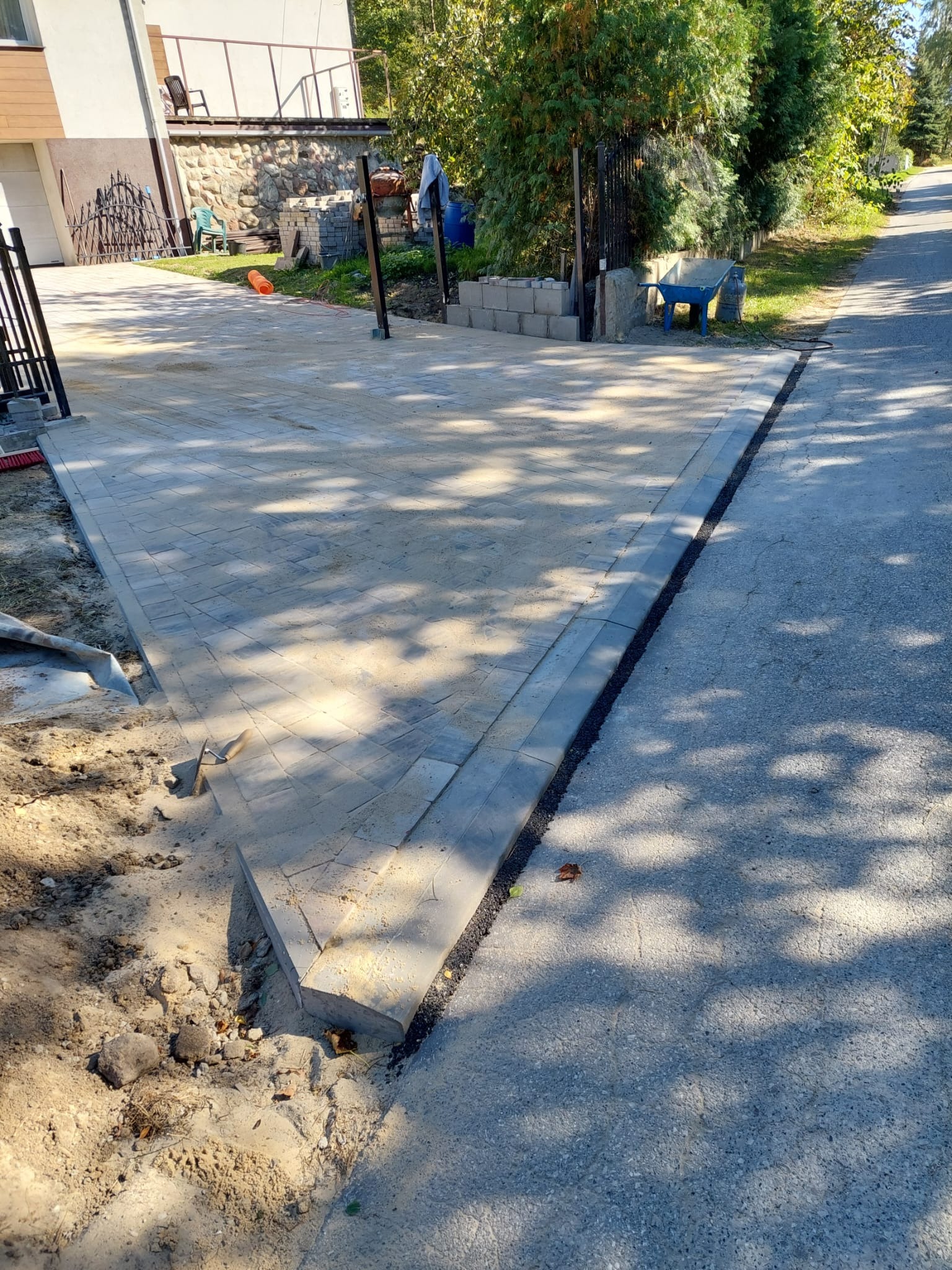 Ułożona kostka brukowa na podjeździe, widoczne narzędzia i piasek. Częściowo wykończona nawierzchnia w cieniu drzew, z widokiem na dom i ulicę.