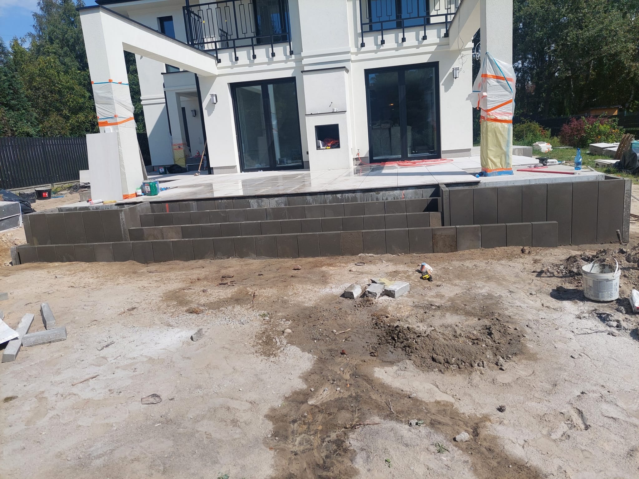 Budowa tarasu z kostki brukowej przy nowoczesnym domu. Widoczne ułożone stopnie z kostki, przygotowany teren i materiały budowlane. Słupy osłonięte folią.