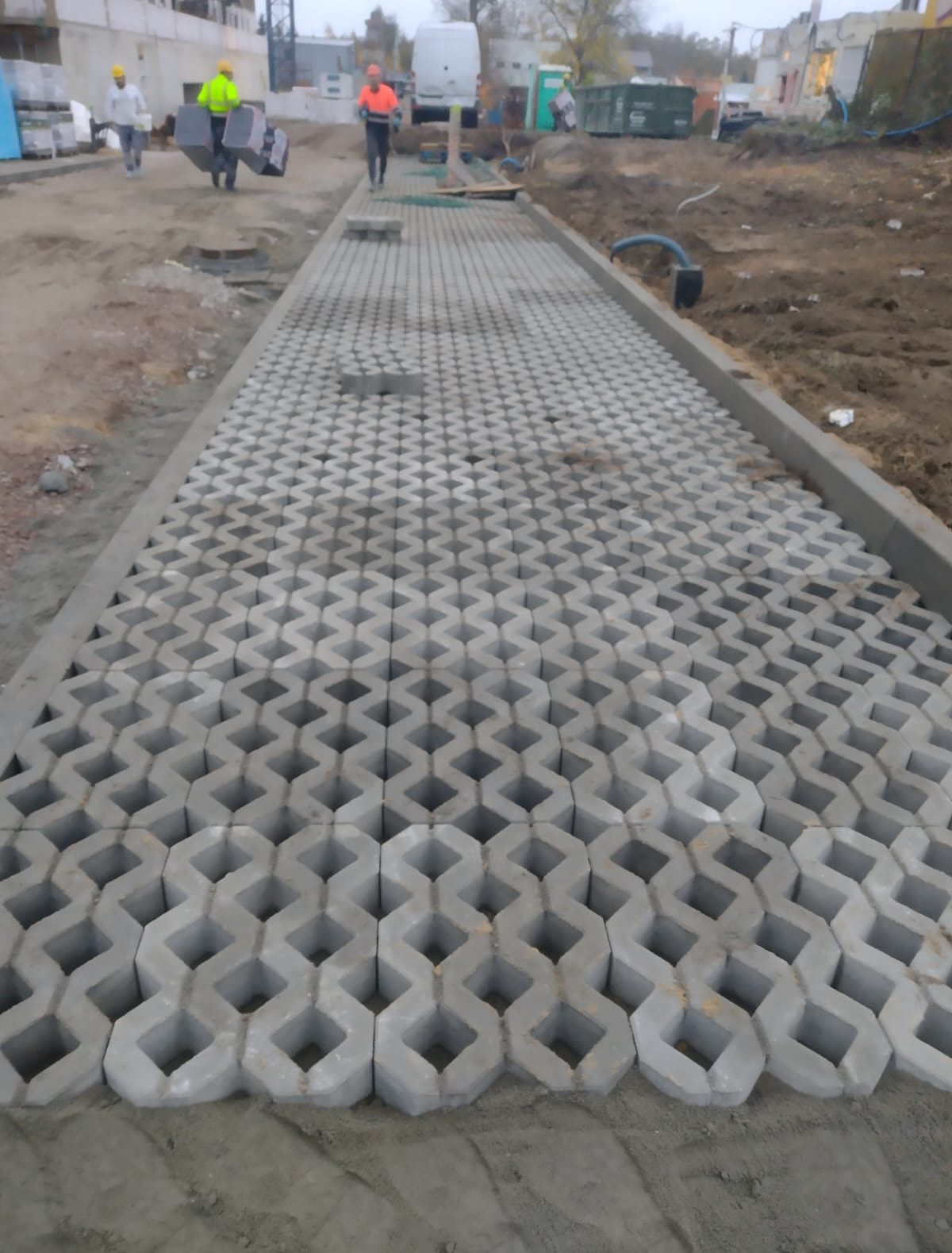 Fragment układania ażurowej kostki brukowej na podbudowie. W tle pracownicy i materiały budowlane.