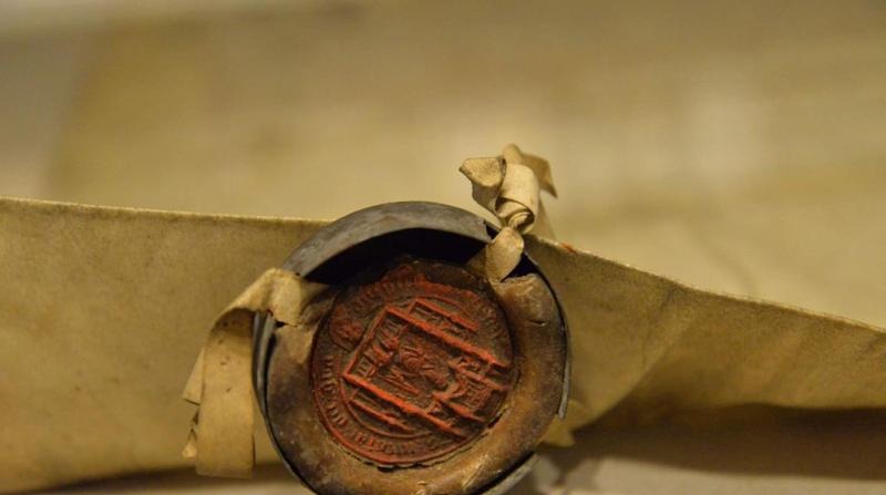 Dokument z pieczęcią lakową, zamknięty metalową obręczą, na beżowym tle. Pieczęć z wyraźnym, czerwonym wzorem herbowym. Detal archiwalny.