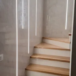 Schody drewniane z podświetleniem LED w ścianie, nowoczesny design. Oświetlenie liniowe podkreśla fakturę ściany. Wykończenie w odcieniach bieli i drewna.