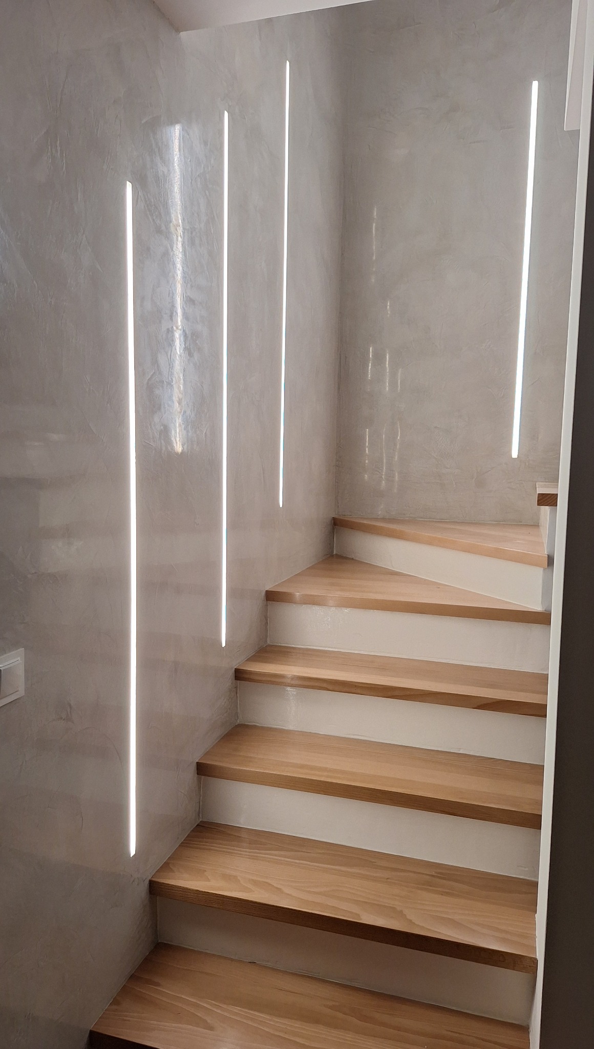 Schody drewniane z podświetleniem LED w ścianie, nowoczesny design. Oświetlenie liniowe podkreśla fakturę ściany. Wykończenie w odcieniach bieli i drewna.