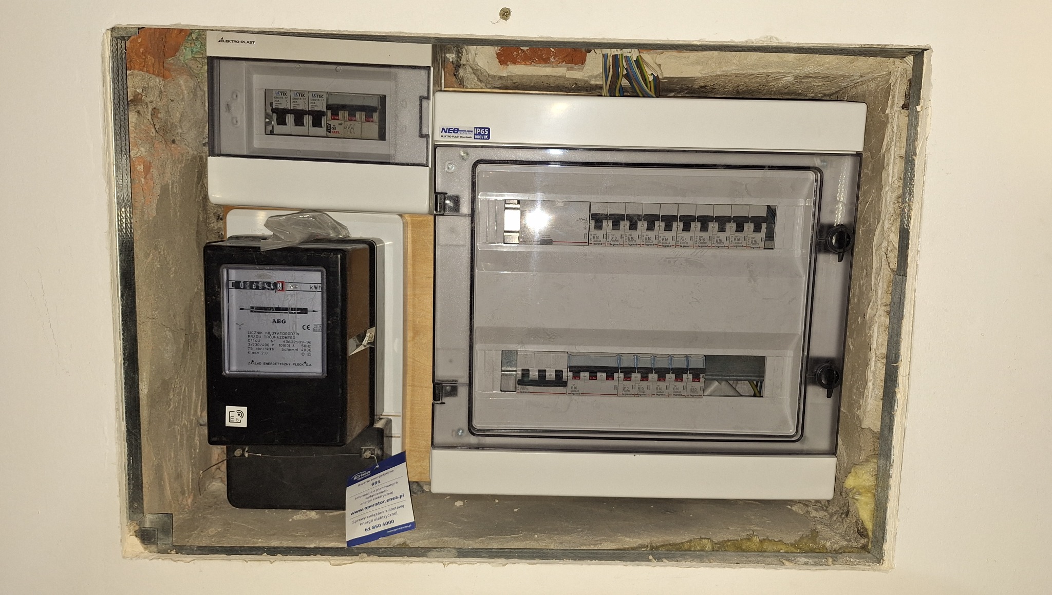 Rozdzielnia elektryczna z licznikiem, zamontowana w ścianie, widoczne zabezpieczenia i przewody. Surowy montaż w otworze ściany. Tabliczka z numerem telefonu.