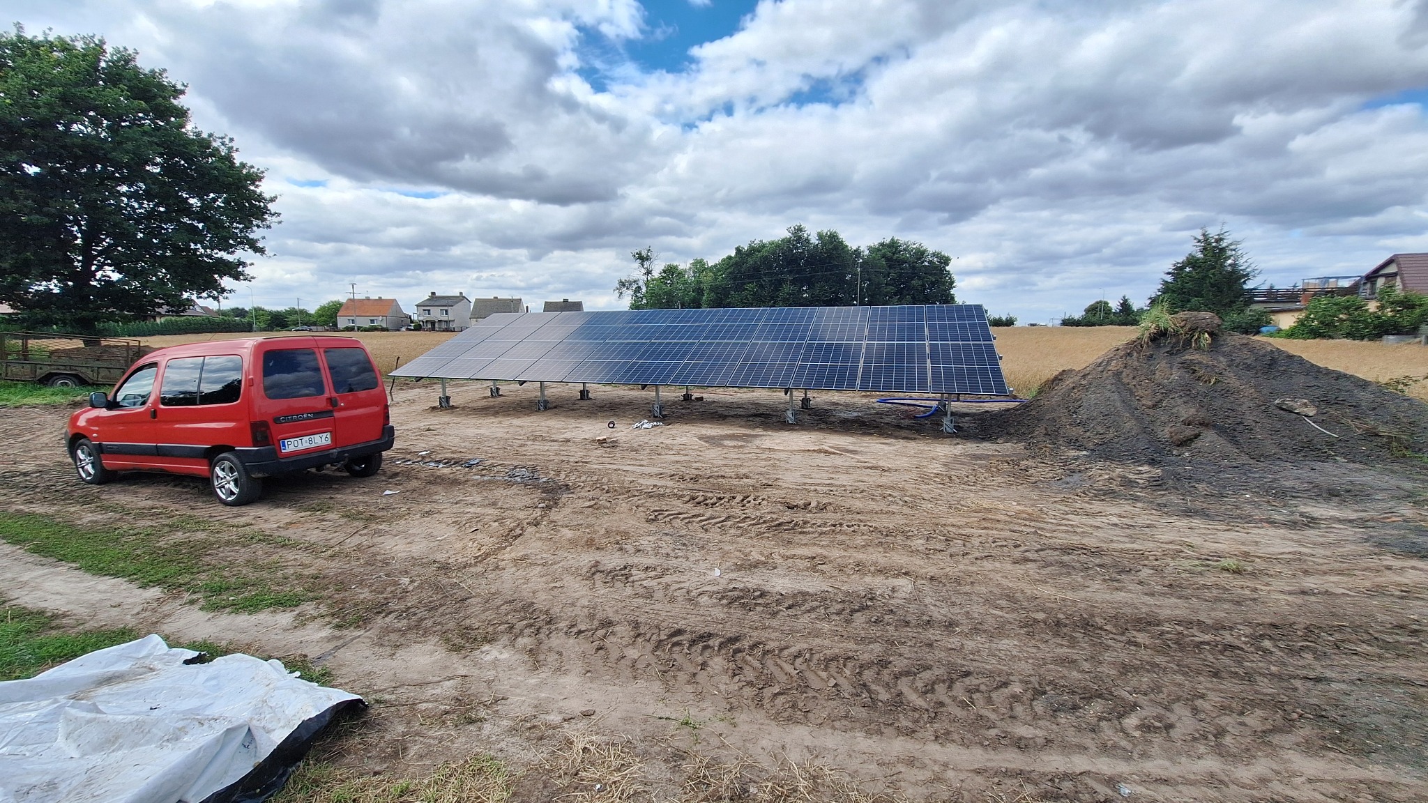 Instalacja paneli fotowoltaicznych na gruncie, widoczny czerwony samochód dostawczy Citroen Berlingo i sterta ziemi. Pochmurne niebo w tle.