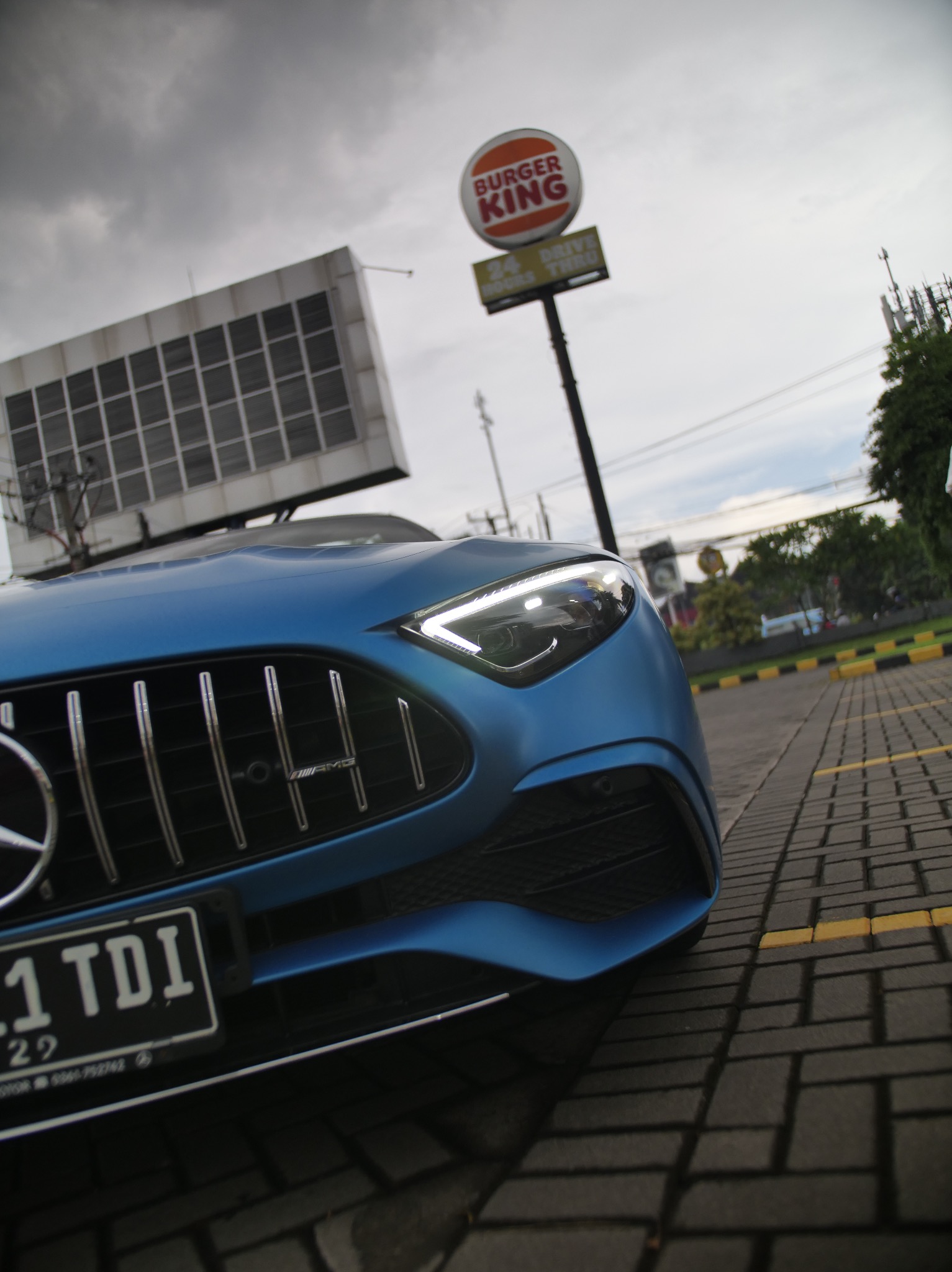 Błękitny Mercedes-AMG z widoczną tablicą rejestracyjną na tle parkingu i znaku Burger King, uchwycony z niskiego kąta.