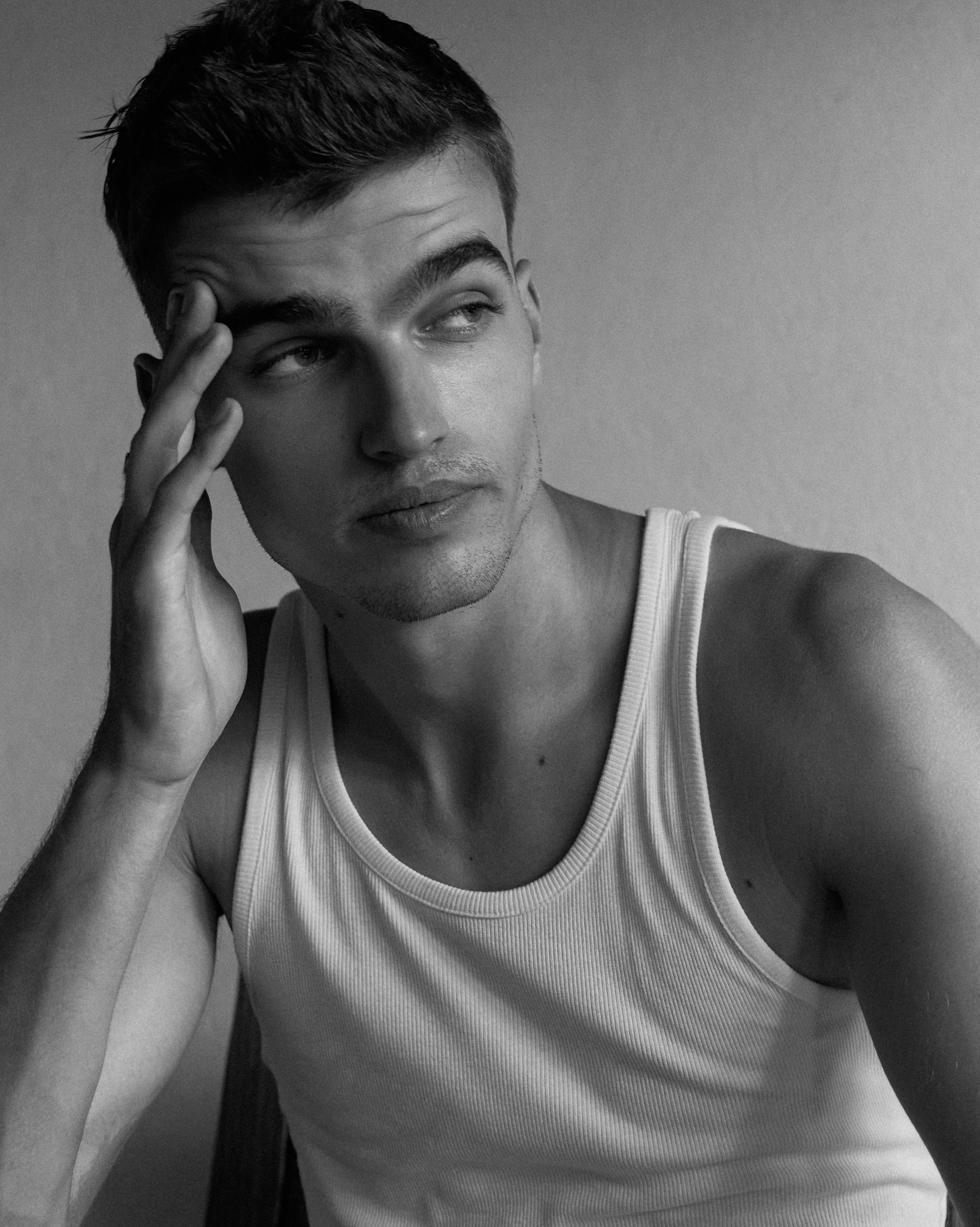 Czarno-biały portret mężczyzny w białym podkoszulku, z ręką przy skroni, spoglądającego w bok. Delikatne światło podkreśla rysy twarzy i muskulaturę.