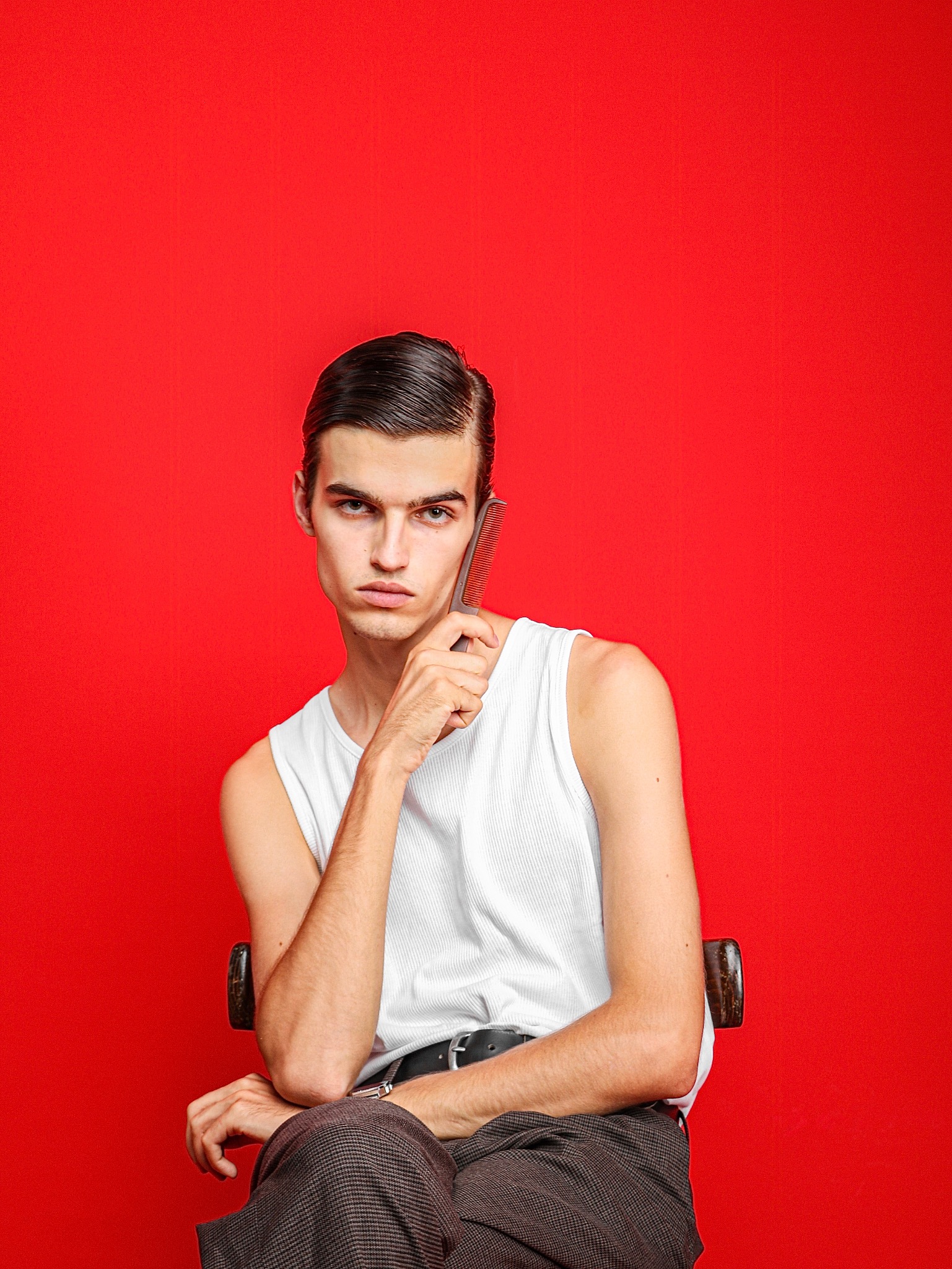 Mężczyzna z gładko zaczesanymi włosami, w białym podkoszulku, pozuje z brązowym grzebieniem na tle intensywnej czerwieni.