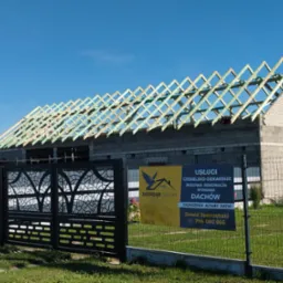 Wznoszona więźba dachowa z impregnacją na budynku w trakcie budowy, widoczny baner reklamowy firmy dekarskiej i ozdobne ogrodzenie posesji.