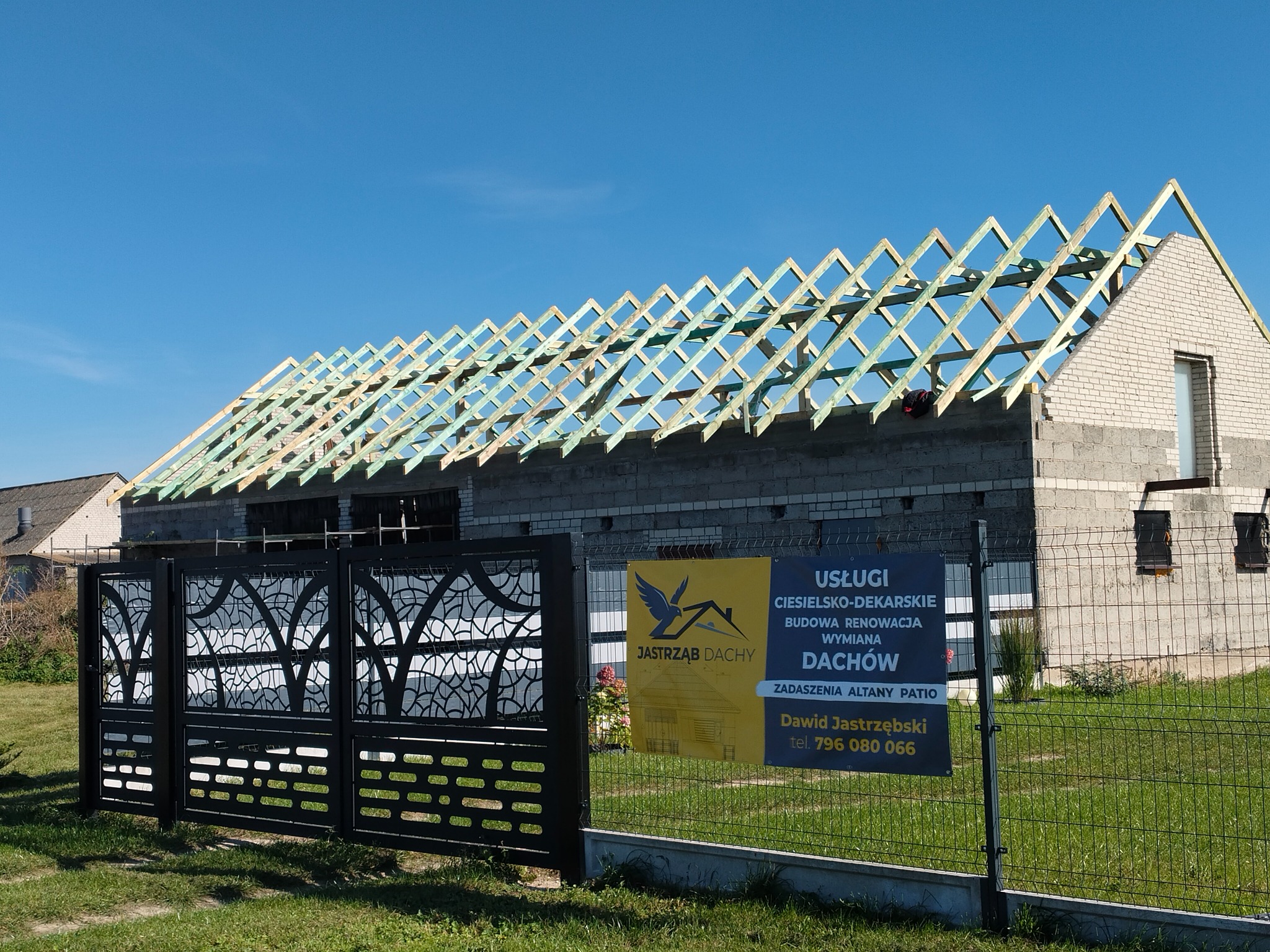 Wznoszona więźba dachowa z impregnacją na budynku w trakcie budowy, widoczny baner reklamowy firmy dekarskiej i ozdobne ogrodzenie posesji.
