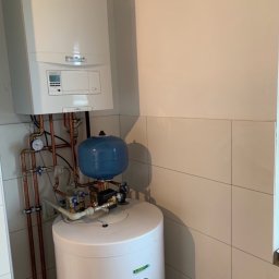 Złota Rączka Kraków - Biały bojler z niebieskim zbiornikiem wyrównawczym i miedzianymi rurami w jasnej kotłowni z kafelkami. Widoczny manometr i zawory.