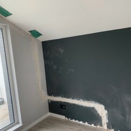 Złota Rączka Kraków - Szpachlowanie ściany w pokoju z oknem. Widoczne gniazdka elektryczne, surowy stan wykończenia. Szara ściana, ciemna ściana i panele podłogowe.