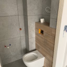 Złota Rączka Kraków - Świeżo wykończona łazienka z szarą marmurową płytką i podwieszaną toaletą na drewnianej obudowie. Widoczne przyłącza wody i kanalizacji.