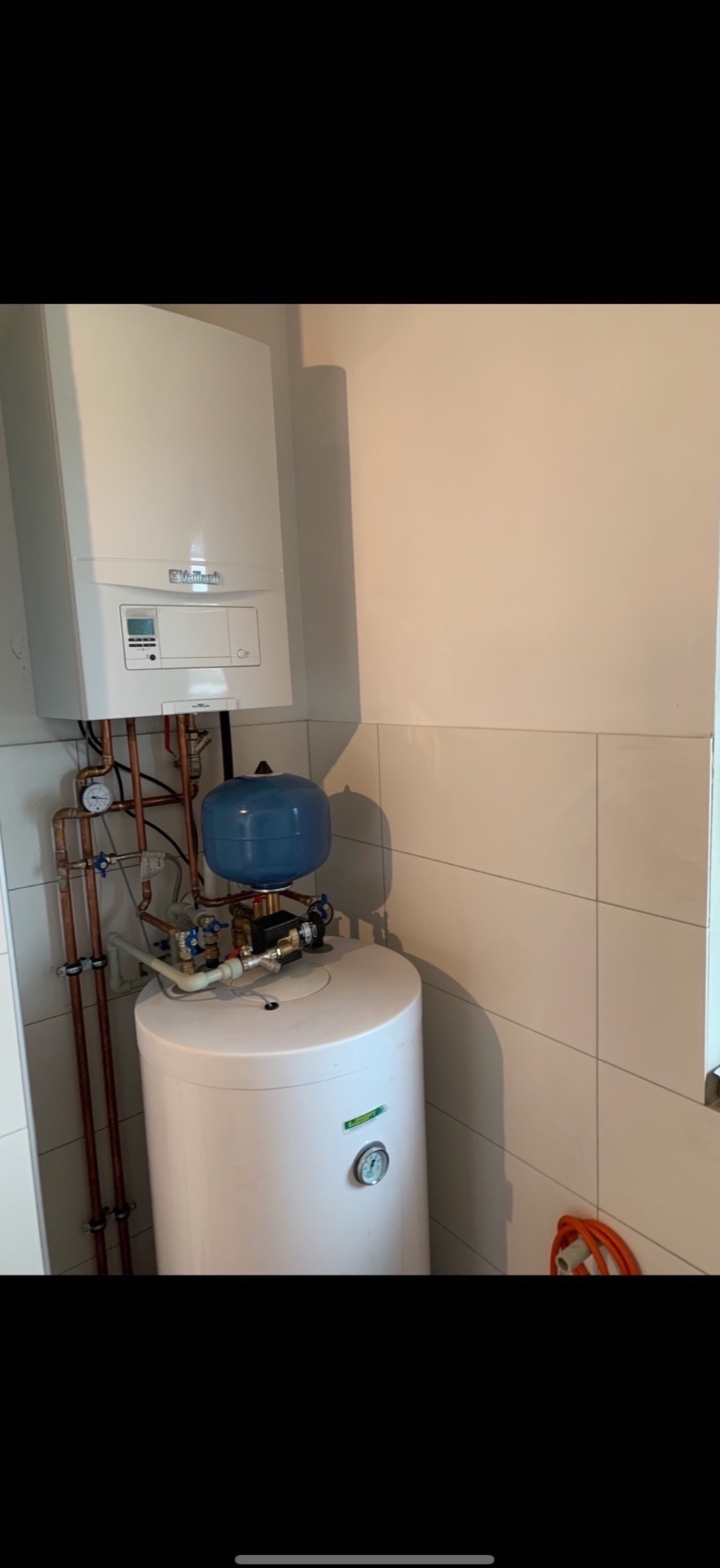 Biały bojler z niebieskim zbiornikiem wyrównawczym i miedzianymi rurami w jasnej kotłowni z kafelkami. Widoczny manometr i zawory.