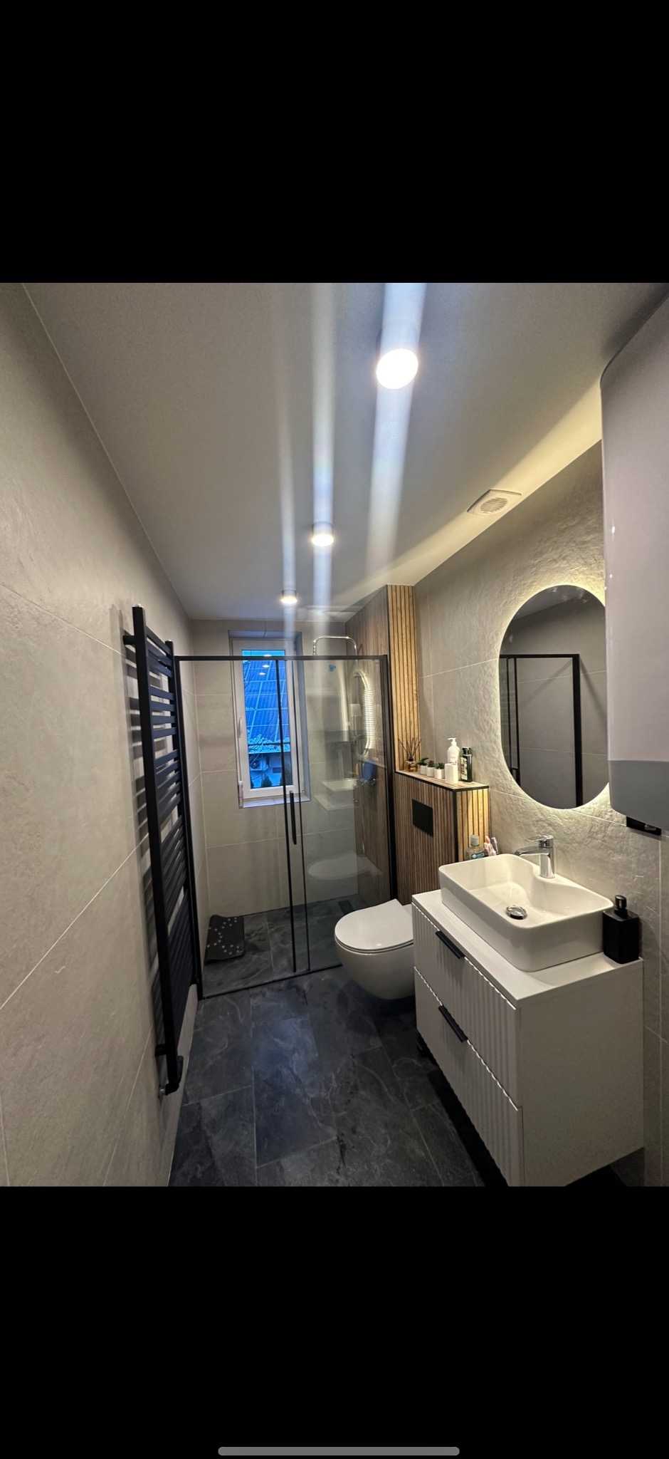 Nowoczesna łazienka z kabiną prysznicową typu walk-in, umywalką na szafce i okrągłym lustrem z podświetleniem LED. Ciemna podłoga i jasne ściany tworzą elegancki kontrast.