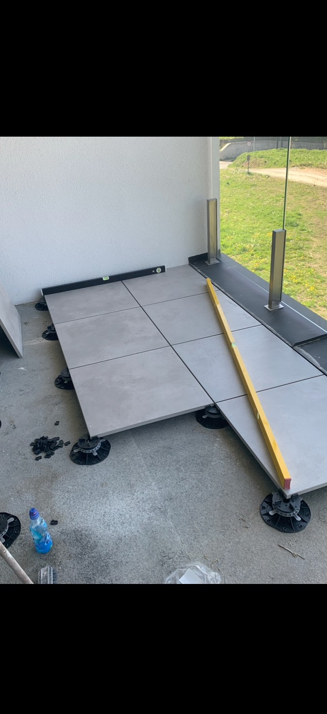 Układanie szarych płytek na regulowanych wspornikach na balkonie z przeszkloną balustradą, poziomica w użyciu. Widoczne narzędzia i butelka wody.