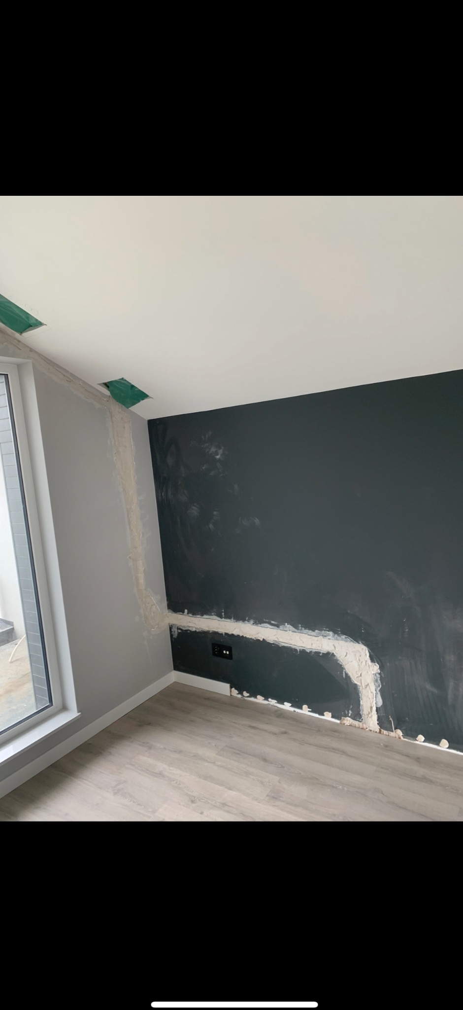 Szpachlowanie ściany w pokoju z oknem. Widoczne gniazdka elektryczne, surowy stan wykończenia. Szara ściana, ciemna ściana i panele podłogowe.