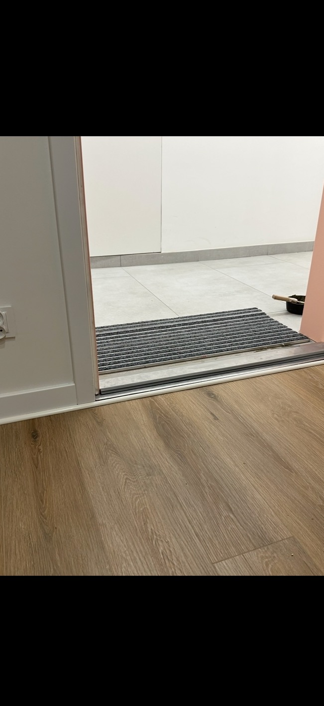 Wykończona podłoga z paneli w odcieniach brązu, widoczny próg i wycieraczka przed wejściem do pomieszczenia z szarymi płytkami. Świeżo po remoncie.