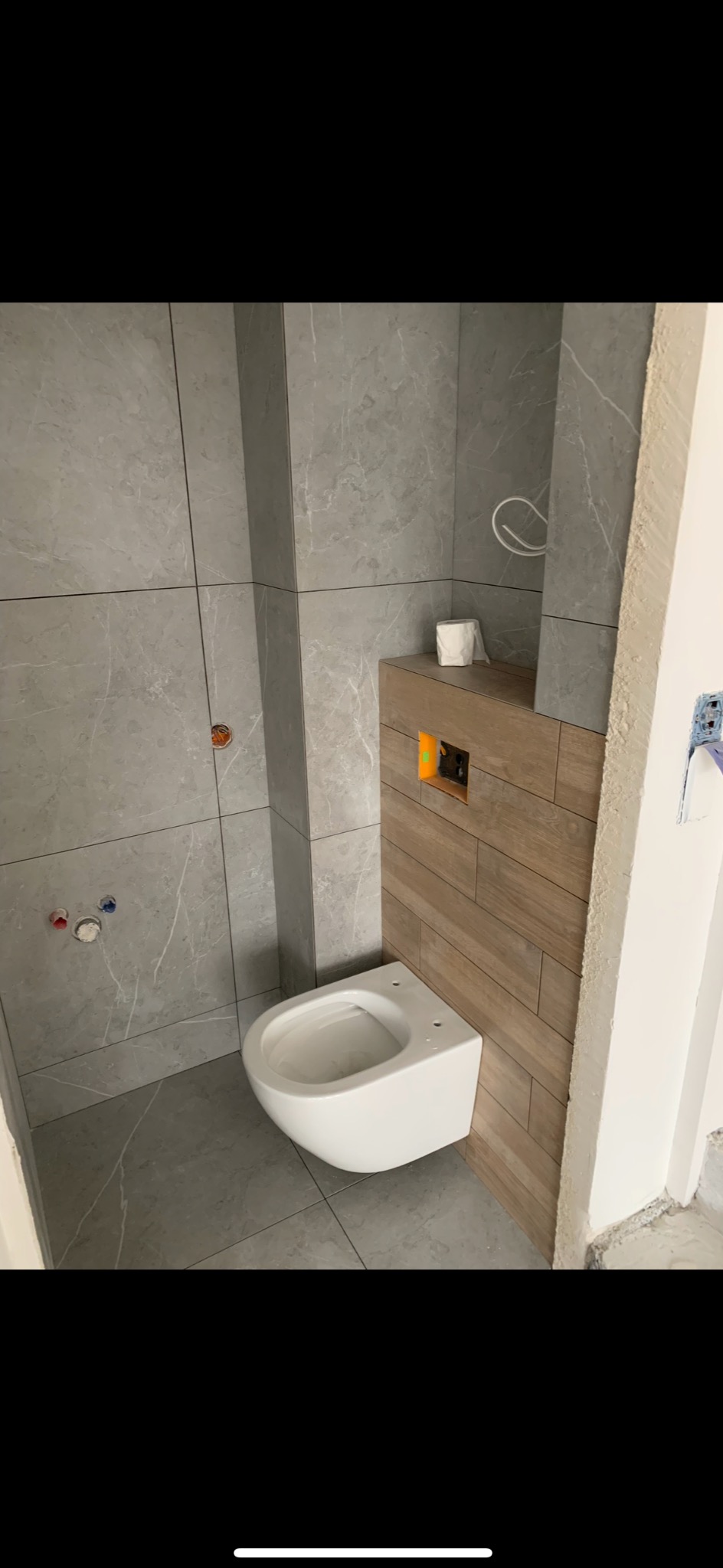 Świeżo wykończona łazienka z szarą marmurową płytką i podwieszaną toaletą na drewnianej obudowie. Widoczne przyłącza wody i kanalizacji.