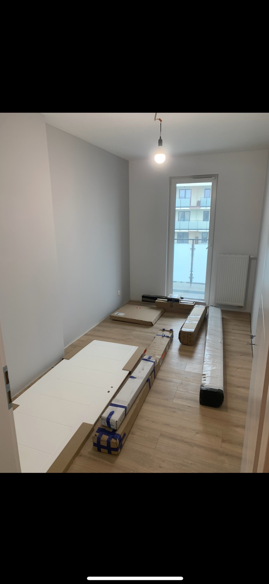 Pokój z parkietem i elementami mebli w kartonach przed montażem. Widoczne białe fronty i zapakowane elementy, surowa żarówka zwisająca z sufitu.