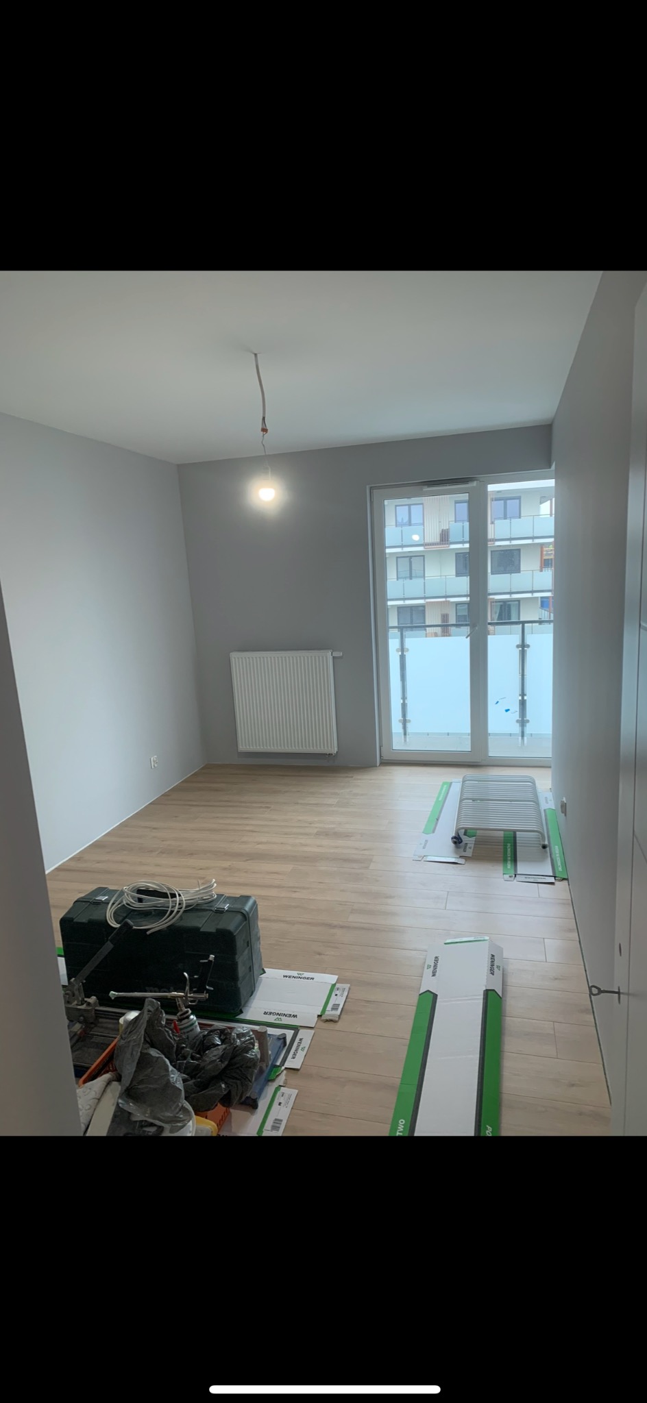 Szare ściany i panele podłogowe w pokoju z balkonem. Widoczny grzejnik, narzędzia i materiały budowlane. Przygotowanie do remontu mieszkania.