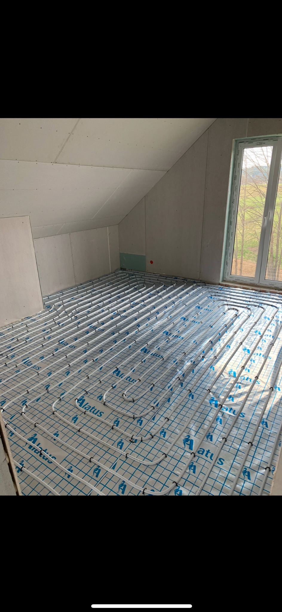 Instalacja ogrzewania podłogowego w surowym wnętrzu poddasza z oknem. Białe rury ułożone na folii z niebieską siatką i logotypem Alatus.