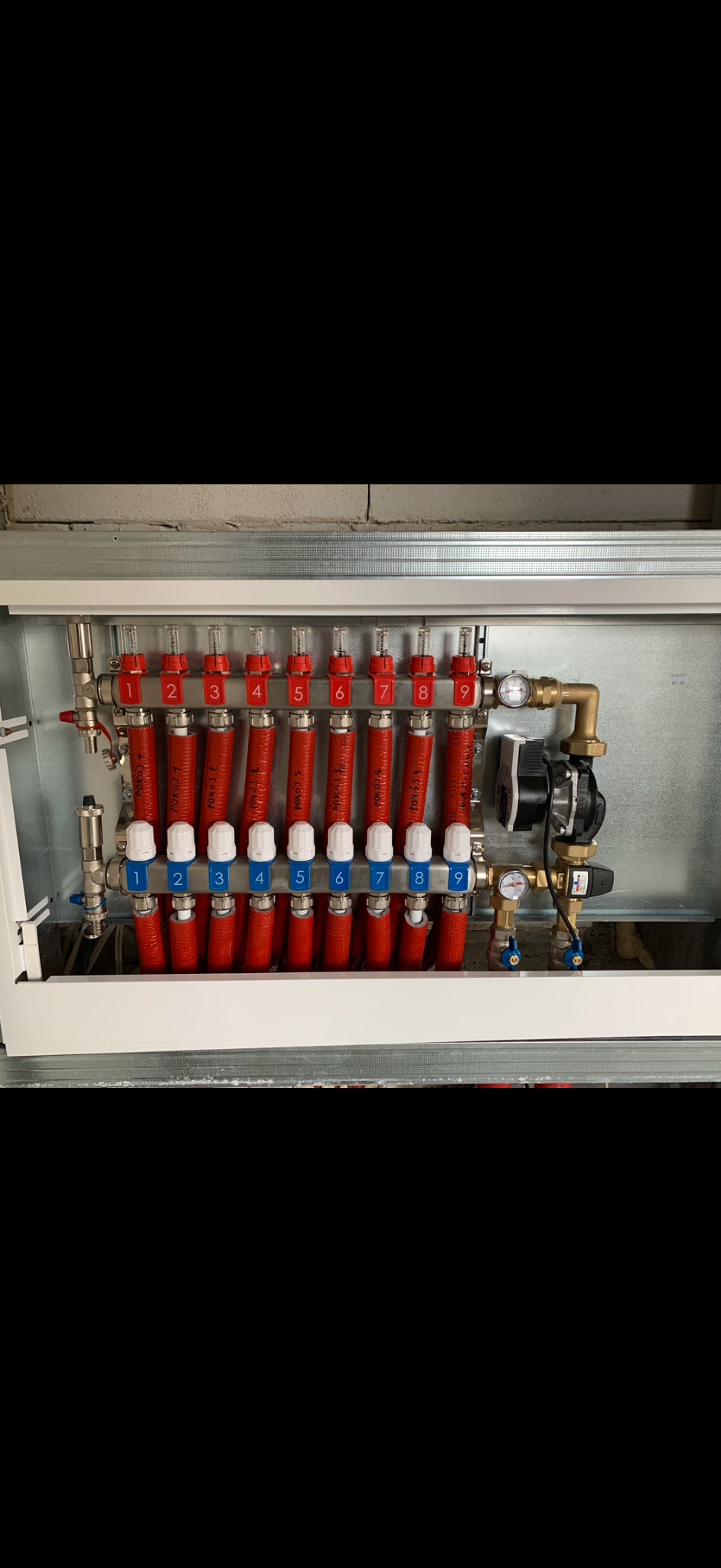 Rozdzielacz ogrzewania podłogowego z czerwonymi rurami oznaczonymi numerami 1-9, z białymi zaworami i manometrem. Metalowa obudowa, widok z przodu.