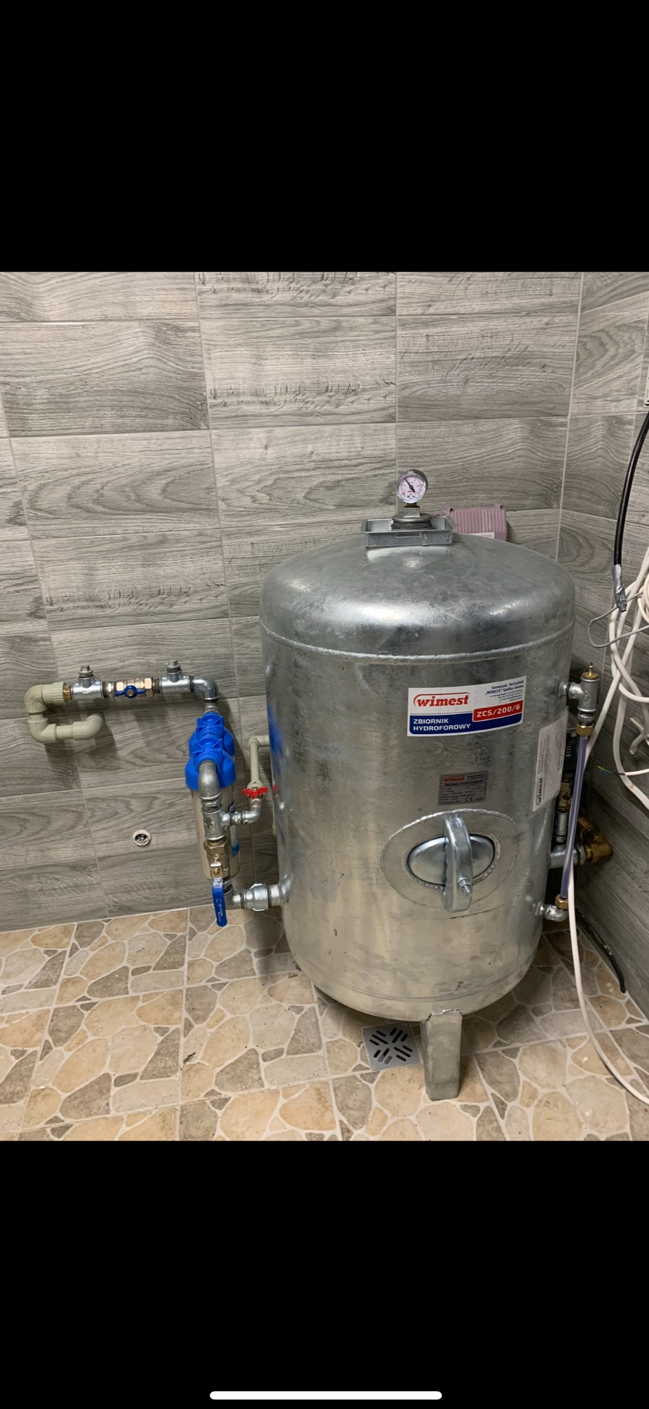 Zamontowany hydrofor WIMEST ZCS/200/6 z manometrem i filtrem wody, widoczne przyłącza hydrauliczne, na tle płytek ściennych imitujących drewno i podłogowych w odcieniach beżu.
