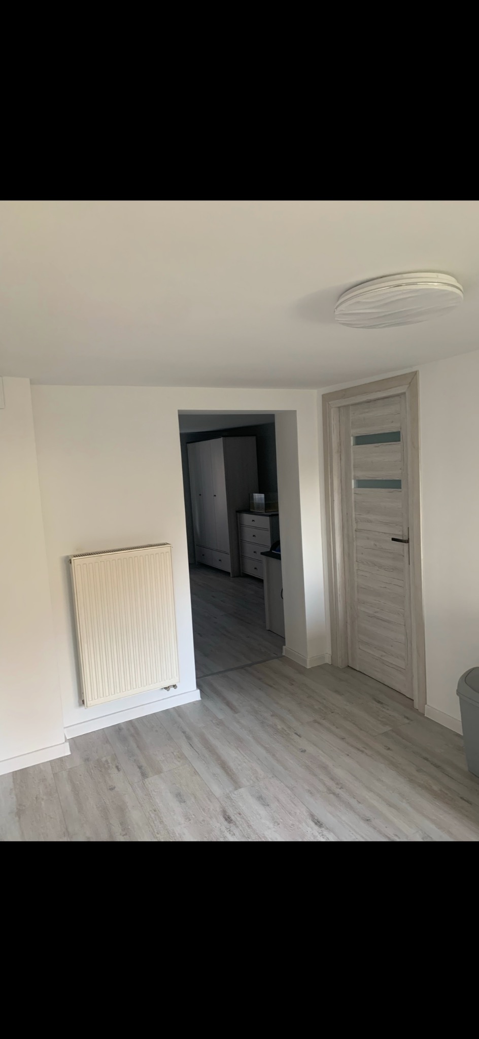 Jasne, odnowione wnętrze z panelami podłogowymi imitującymi drewno, białą ścianą z grzejnikiem i drzwiami z szarymi wstawkami. Widok na kolejne pomieszczenie.