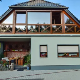 Elewacja domu z oknami w drewnianych ramach i roletami w Nysie. Górne okna o nietypowym kształcie. Na parapecie skrzynki z kwiatami. Widok z zewnątrz.