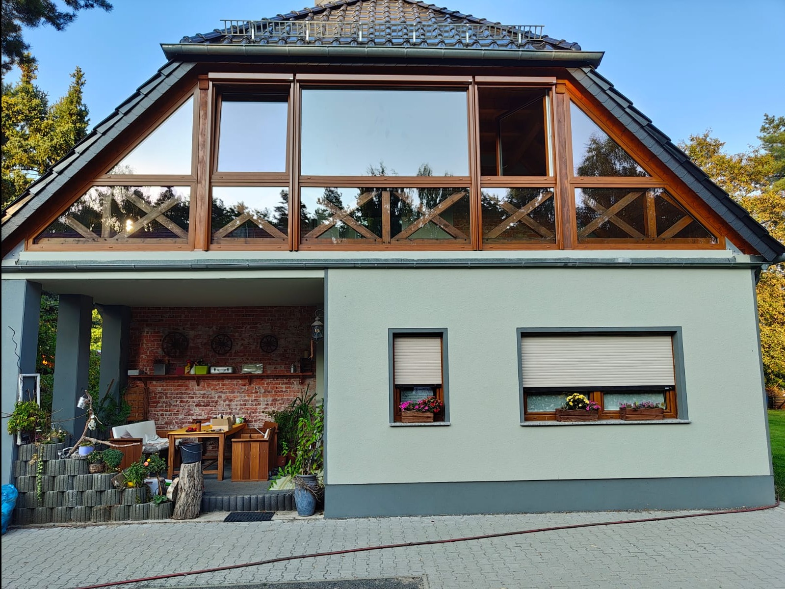 Elewacja domu z oknami w drewnianych ramach i roletami w Nysie. Górne okna o nietypowym kształcie. Na parapecie skrzynki z kwiatami. Widok z zewnątrz.