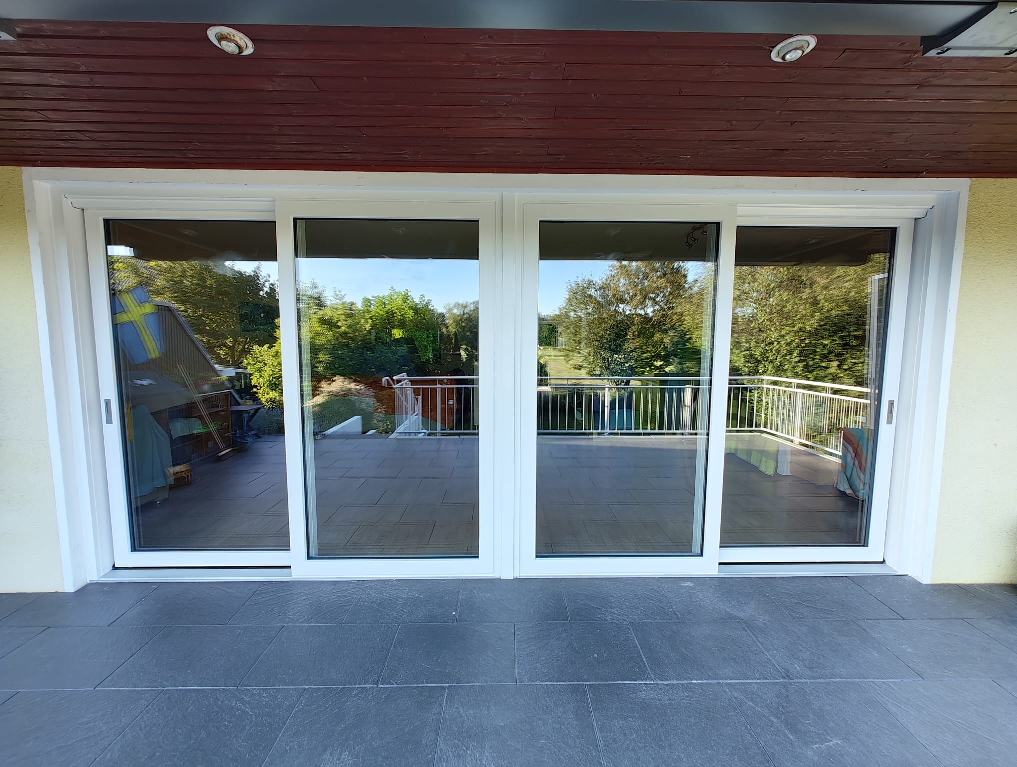 Białe, czteroskrzydłowe drzwi przesuwne z widokiem na balkon i zieleń. Widoczne szare płytki podłogowe i drewniane wykończenie sufitu.