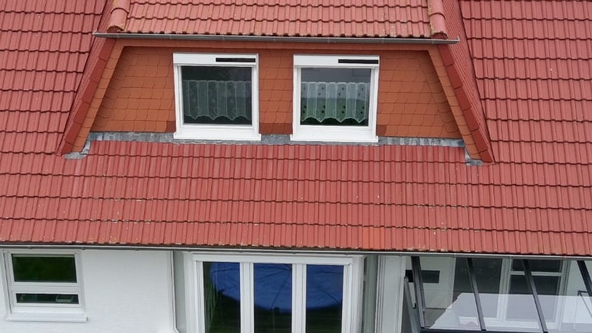 Czerwony dach z oknami dachowymi i częściowo zasłoniętymi roletami. Widoczne firanki w oknach. Ujęcie z góry na dom z białymi ścianami i oknami.