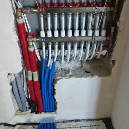 Instal System Patryk Maniek - Rozdzielacz ogrzewania podłogowego z rurami w ścianie i na podłodze, widoczne elementy instalacji w trakcie montażu, izolacja podłogowa z siatką.