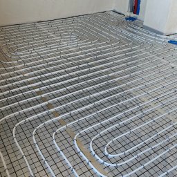 Instal System Patryk Maniek - Białe rury ogrzewania podłogowego ułożone na czarnej siatce w jasnym pomieszczeniu, widoczne niebieskie elementy instalacji przy ścianie.
