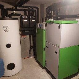 Instal System Patryk Maniek - Nowoczesna kotłownia z białym zasobnikiem Kospel, zielono-szarym piecem SAS i czarnymi rurami instalacji grzewczej w pomieszczeniu z szarą płytką na ścianie.