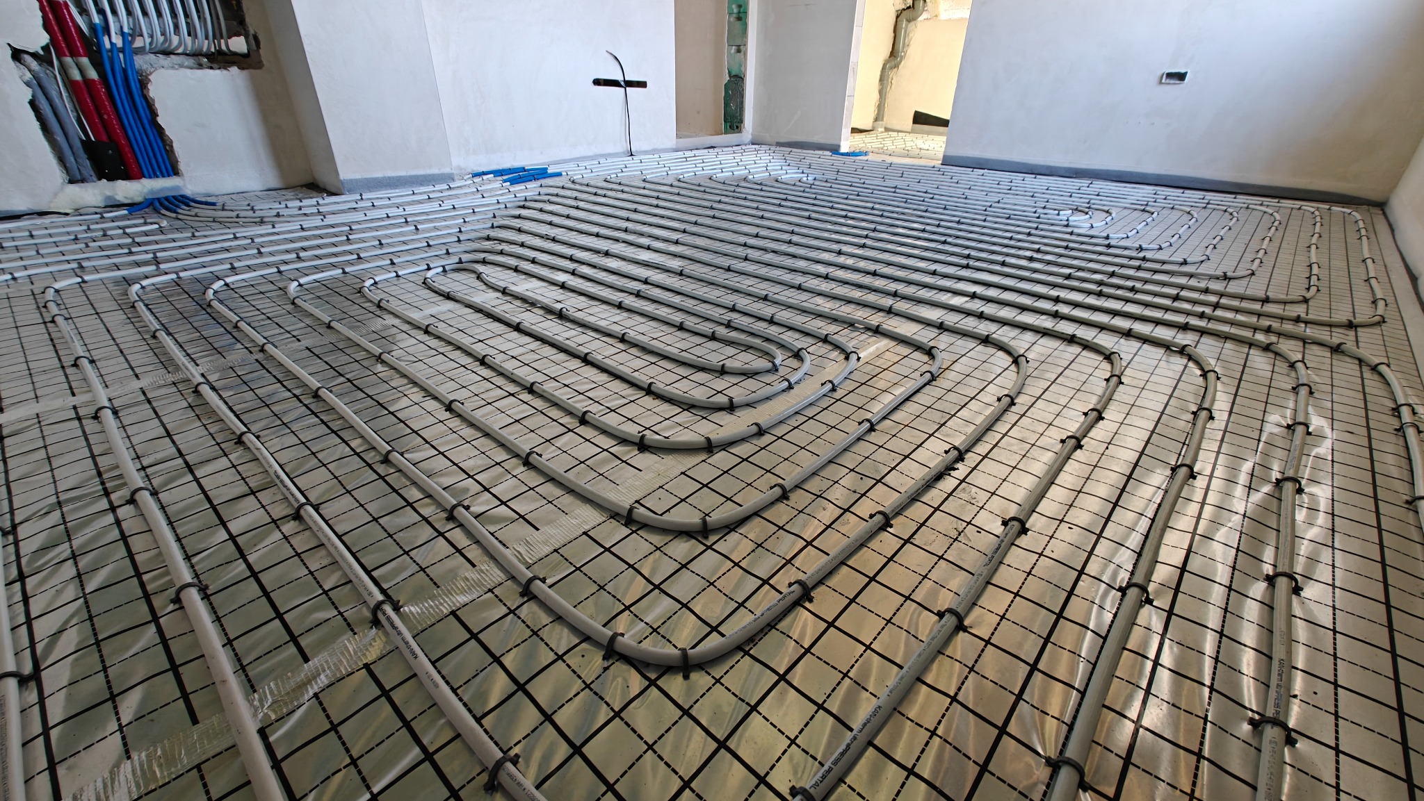 Ułożone rury ogrzewania podłogowego na folii z rastrem, widoczne niebieskie i czerwone rury w ścianie. Instalacja w jasnym pomieszczeniu przed wylaniem posadzki.