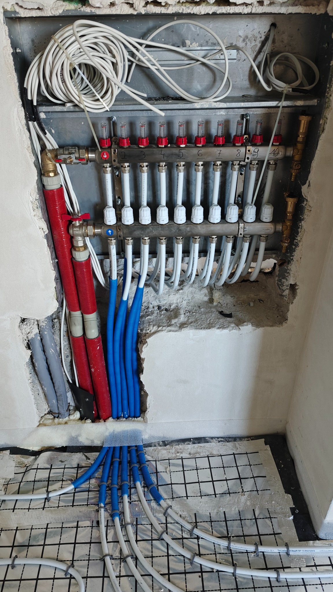 Rozdzielacz ogrzewania podłogowego z rurami w ścianie i na podłodze, widoczne elementy instalacji w trakcie montażu, izolacja podłogowa z siatką.