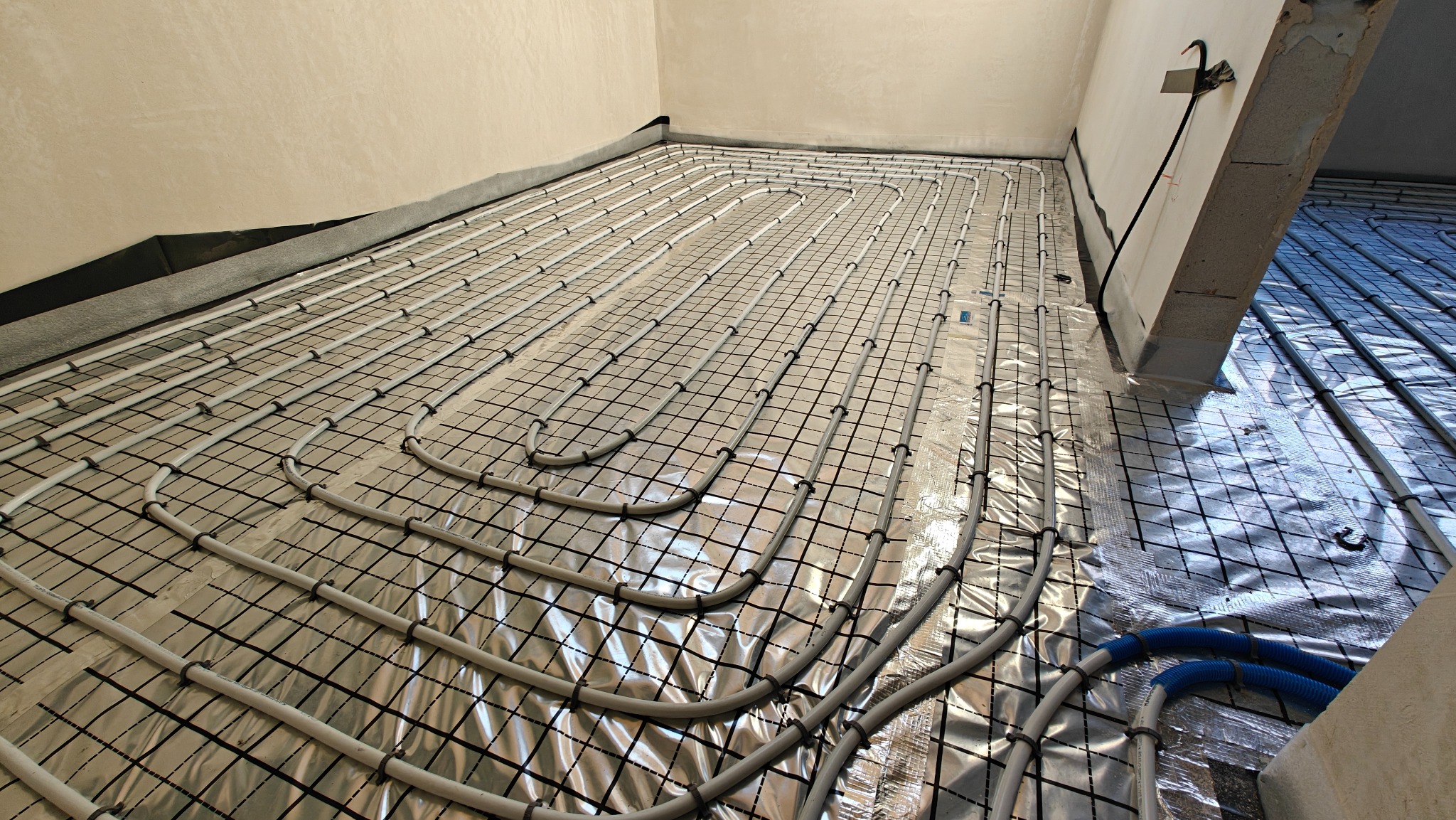 Ułożona instalacja ogrzewania podłogowego na folii aluminiowej z czarną siatką, rury mocowane plastikowymi klipsami, widoczne niebieskie rury doprowadzające.