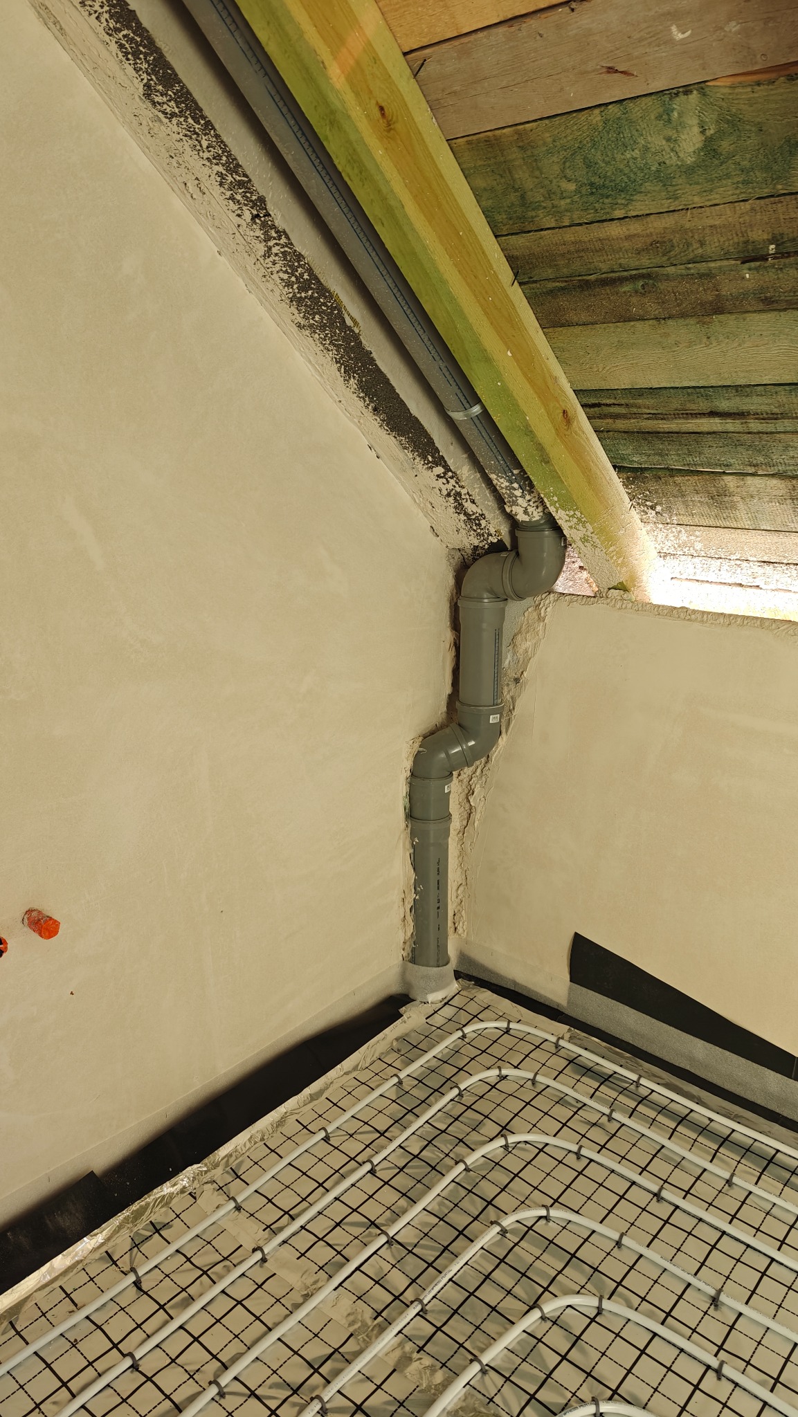 Szare rury instalacji w rogu pomieszczenia z ogrzewaniem podłogowym na folii, widoczne drewniane elementy konstrukcji dachu i fragment ściany w surowym stanie.
