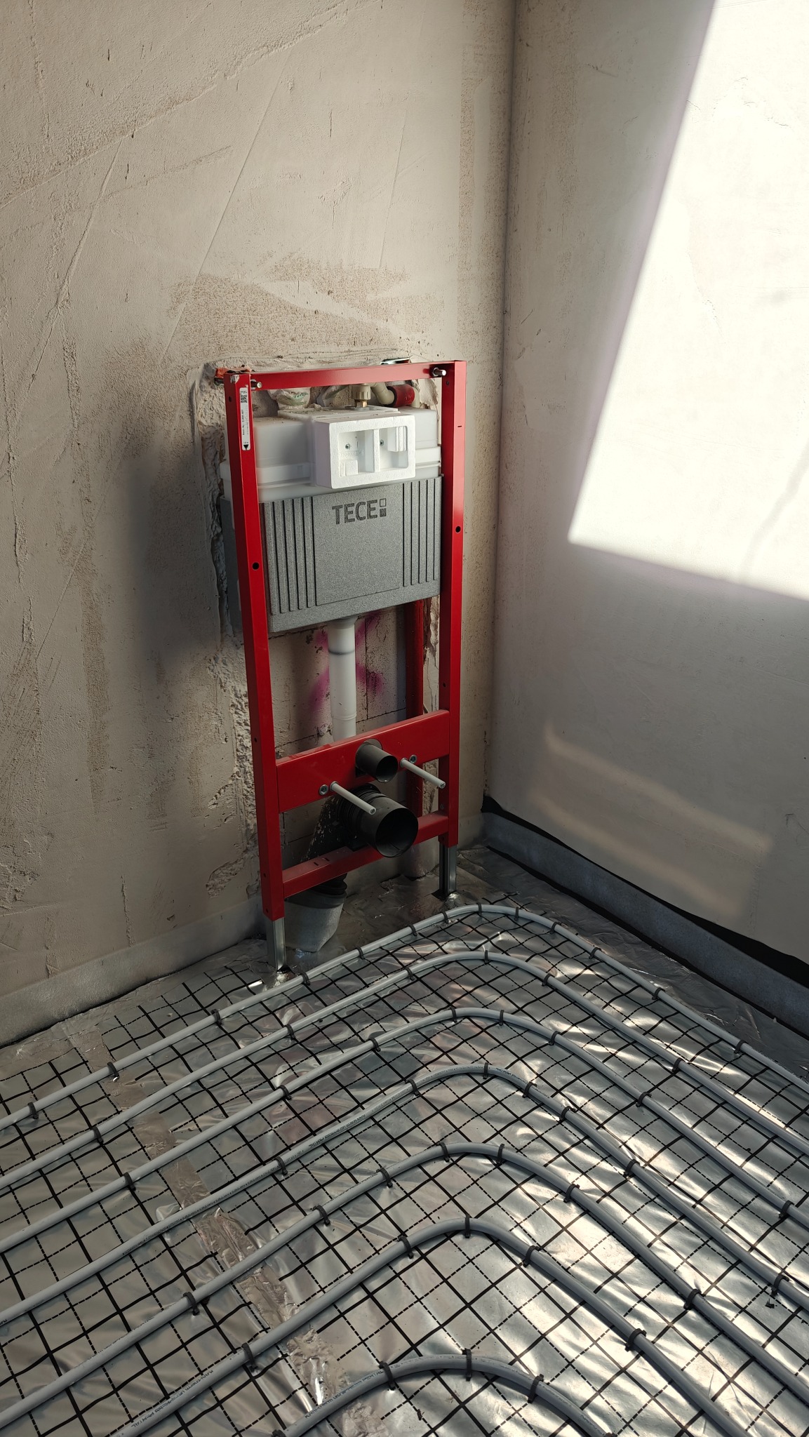 Stelaż podtynkowy TECE z instalacją ogrzewania podłogowego w surowym wnętrzu. Widoczne rury grzewcze na folii izolacyjnej i fragment ściany z niedokończonym tynkiem.