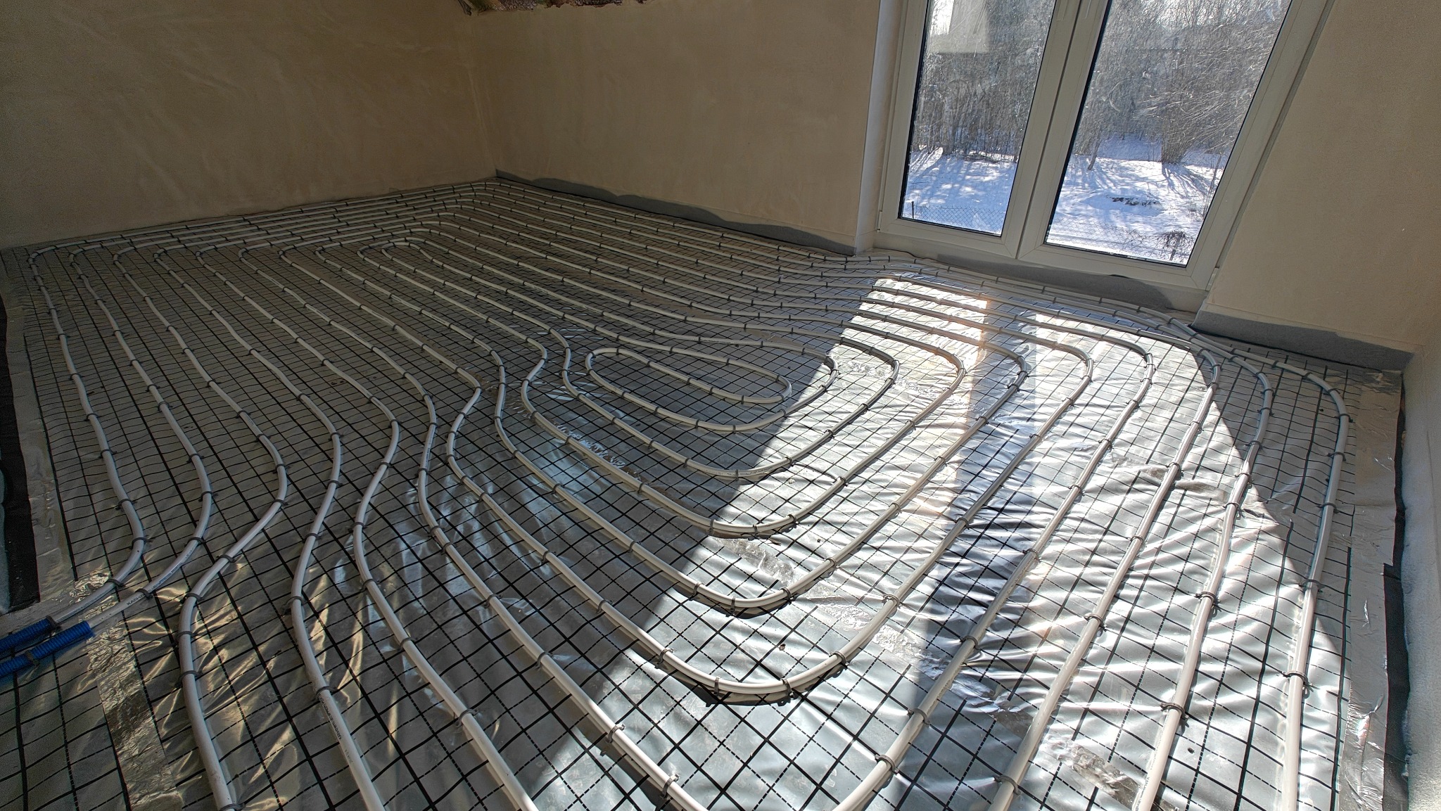 Białe rury ogrzewania podłogowego ułożone na czarnej macie w jasnym pomieszczeniu z oknem. Widoczne promienie słoneczne. Etap instalacji przed wylaniem posadzki.